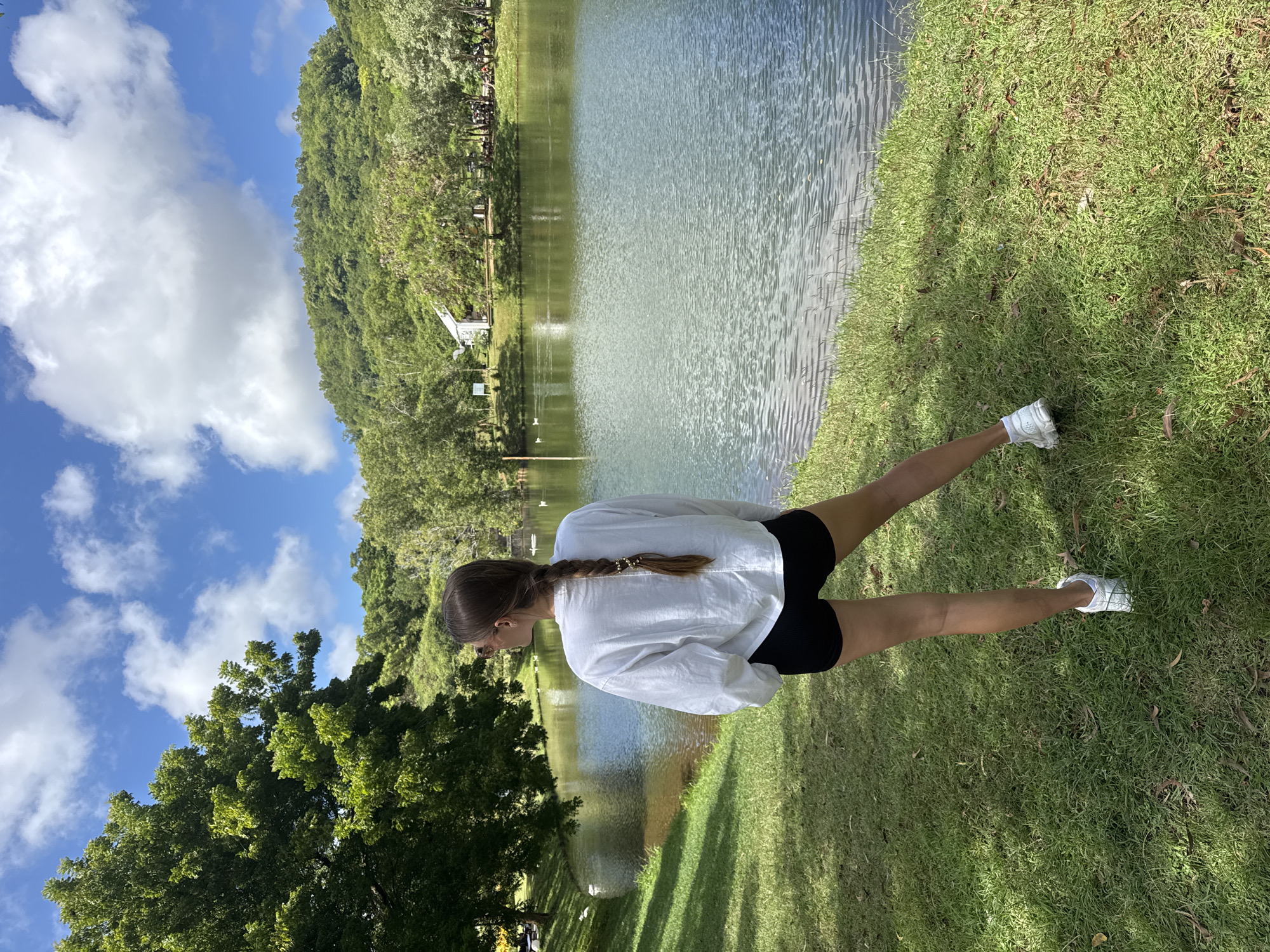 A medium shot depicts a light-skinned young woman from the back, positioned on a grassy bank next to a body of water, possibly a lake or a pond. The location is Unknown, Unknown. She is wearing a white long-sleeved shirt and black shorts with white sneakers. Her brown hair is styled in a single braid that extends down her back. The water surface is slightly rippled, reflecting the sky and surrounding greenery. Behind the water, a dense forest climbs up a hill, and the sky shows scattered white clouds. The immediate foreground is a well-maintained green lawn. The scene appears to be captured on a bright, sunny day.