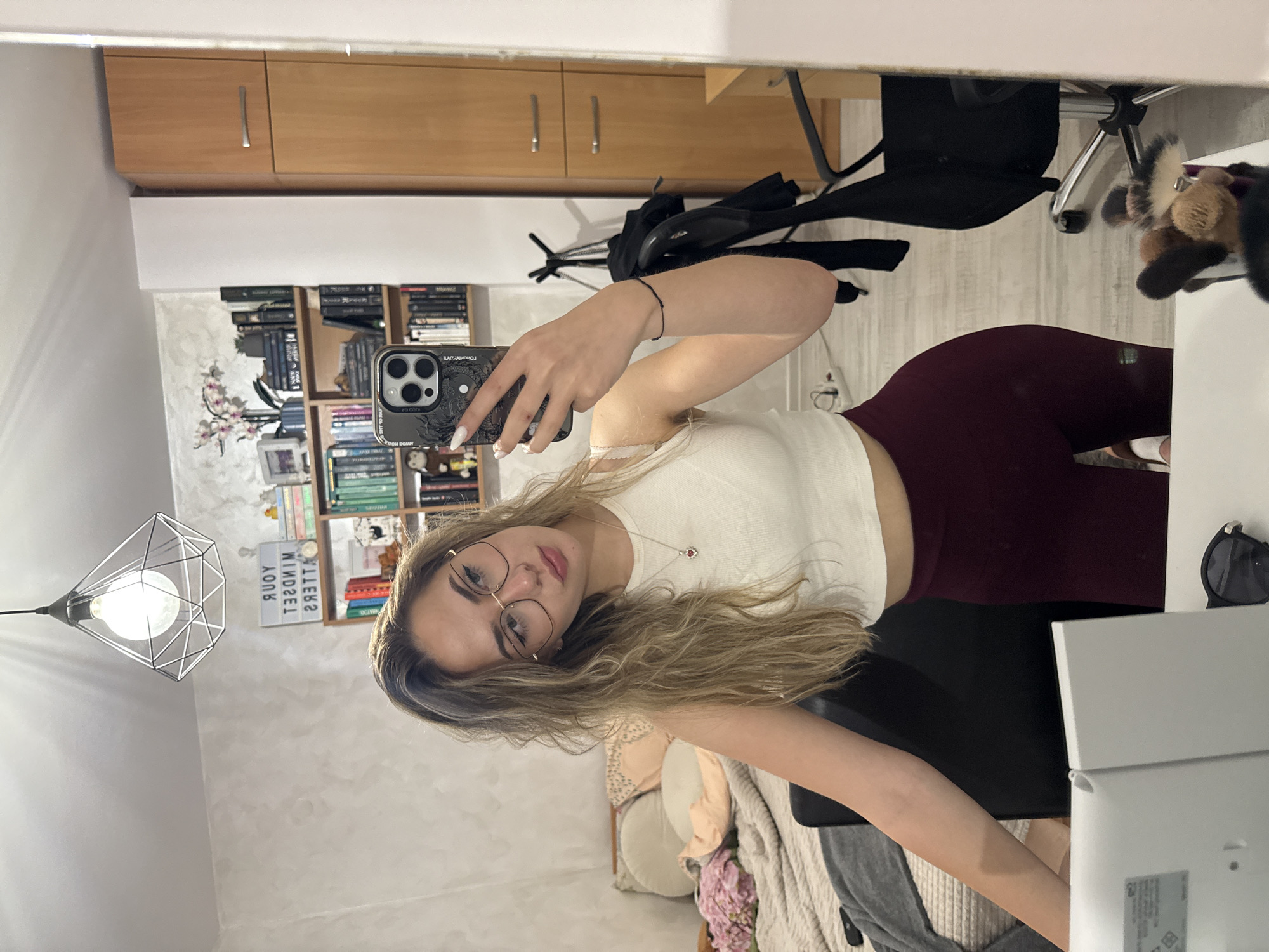 A young woman is taking a selfie in a room with a mirror. She is wearing glasses, a white tank top, and burgundy leggings. The room contains a bookshelf, a desk, a chair, a lamp, and storage cabinets. The location appears to be a residential room, though a specific location in Blagoevgrad, Bulgaria, cannot be determined from the image.