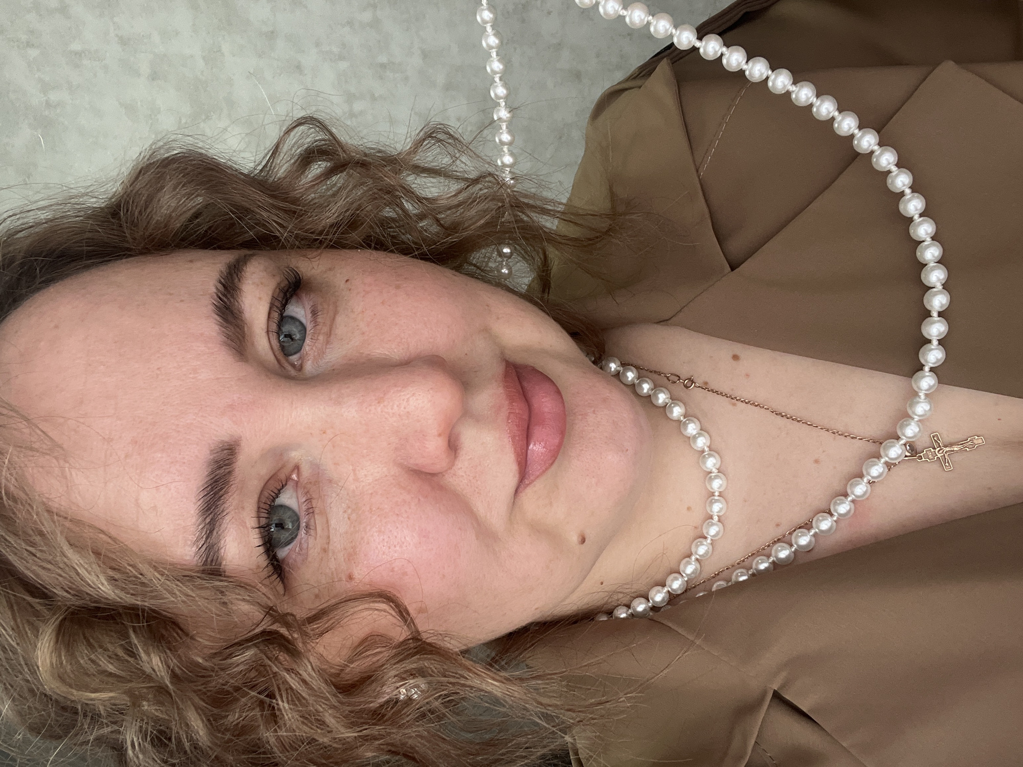 A woman with curly brown hair, fair skin, and light eyes is centered in the image, facing the camera. She is wearing a tan or brown blazer-like garment, and two pearl necklaces, one with a small cross pendant. The background is a blurred light gray. The image is taken indoors. There is no verifiable information to locate it in the city of Chaikovsky, Russia.