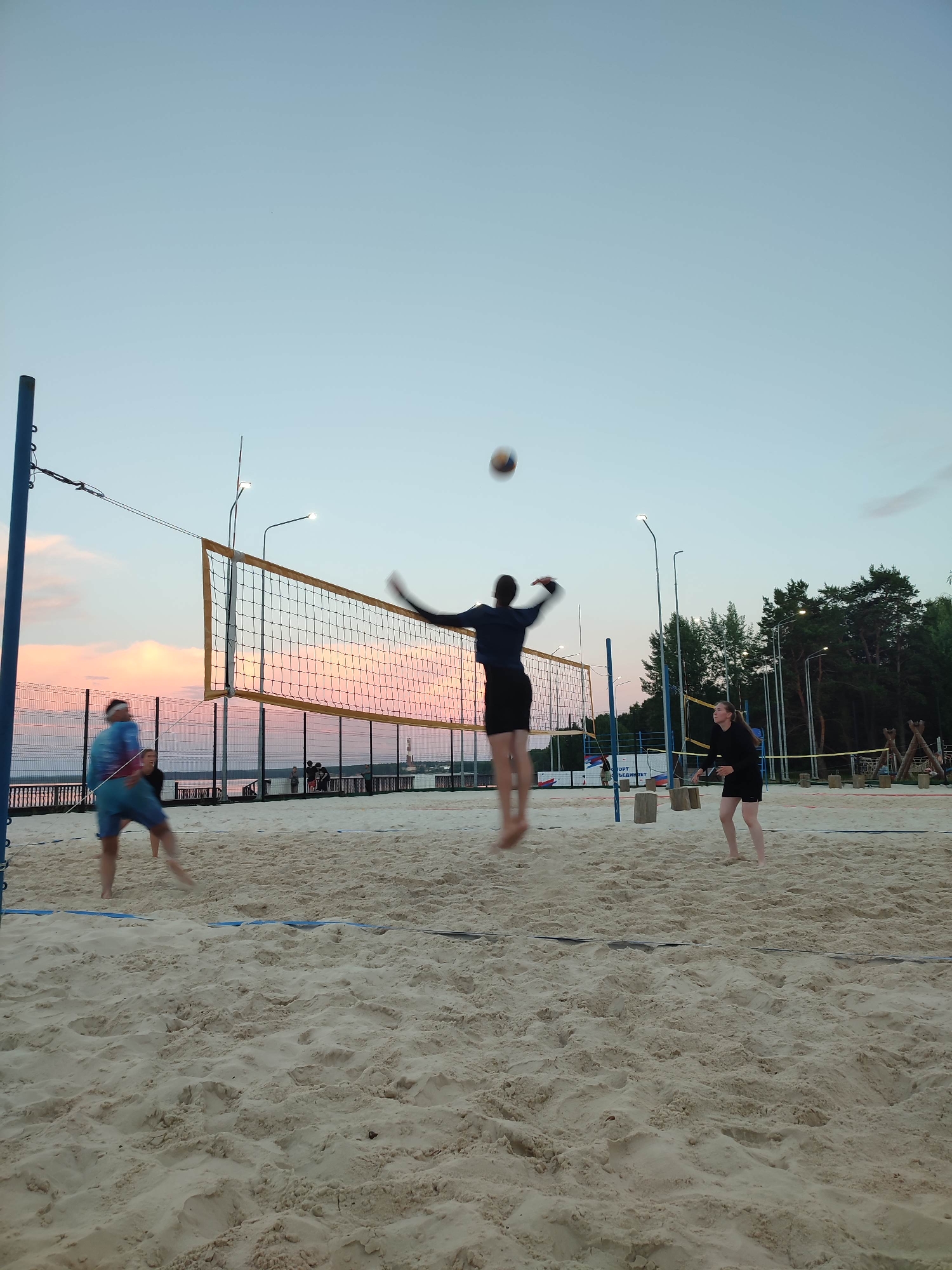 An outdoor volleyball court at dusk features three people playing. The court is made of light-colored sand, with a net strung between two poles. In the center of the image, a person in dark clothing jumps to hit a volleyball, which is visible in the air above them. To the left, a person wearing a colorful shirt and blue shorts is positioned to receive the ball. To the right, a person in black shorts and a black shirt stands ready. The background shows a calm body of water, a distant cityscape with lights, and a line of trees against a pink and blue twilight sky. Streetlights are positioned around the court.