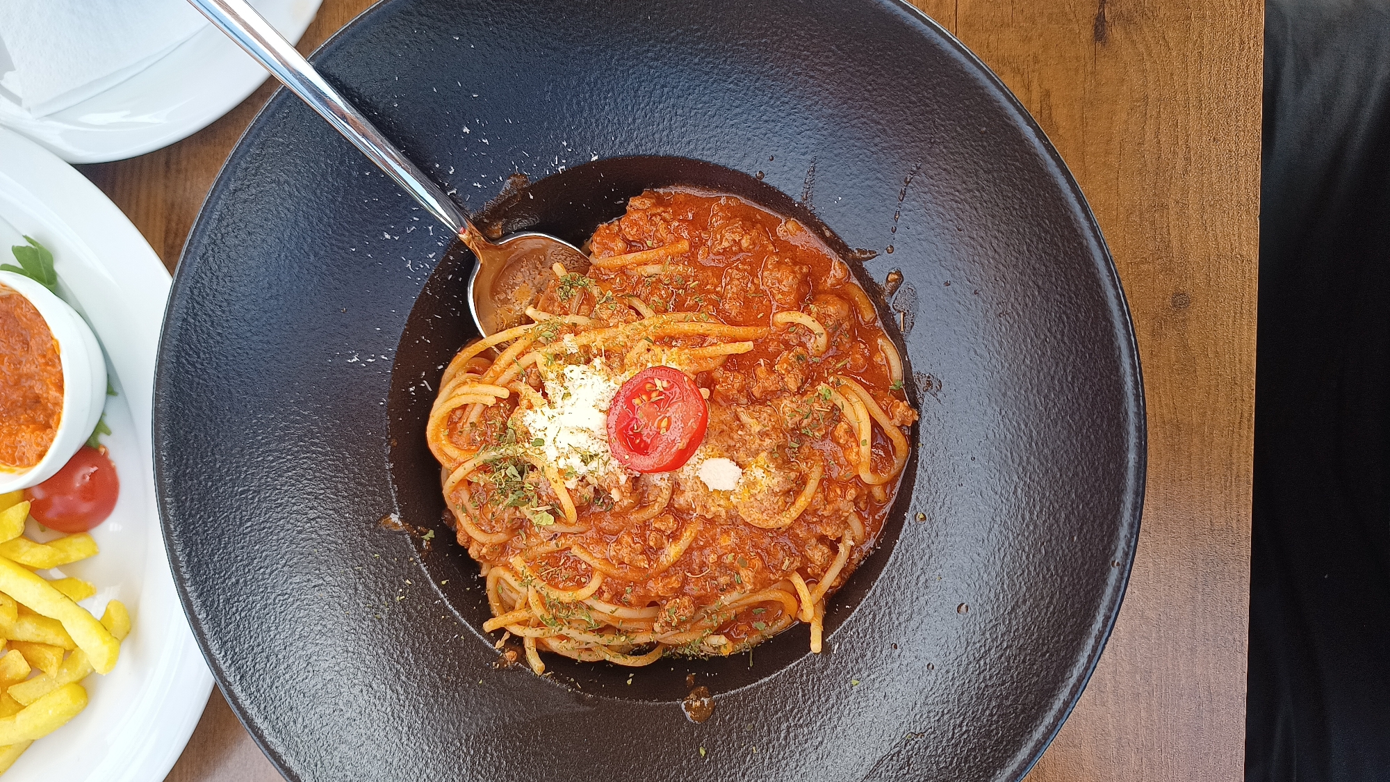 This overhead shot features a deep, textured, dark-colored bowl filled with spaghetti bolognese, centrally placed on a wooden table. The spaghetti is generously coated in a rich, reddish-brown meat sauce. It is garnished with a sprinkle of grated cheese, green dried herbs (likely parsley or oregano), and a single halved cherry tomato positioned on top of the cheese. A silver spoon is resting in the spaghetti, partially submerged in the sauce, with its handle extending towards the upper left.
To the left of the main dish, parts of other accompanying items are visible on white plates. These include a small white ramekin containing a red dip or sauce, several golden-yellow french fries, and another halved cherry tomato. A stack of white plates or napkins is also partially visible in the upper left corner. The table surface is made of warm-toned wood, showing visible grain. The bright, natural lighting suggests the picture was taken during the daytime, possibly in an outdoor or well-lit indoor restaurant setting in Umag, Croatia. No people are visible, and there is no discernible text in the image. The scene depicts a meal ready to be enjoyed.