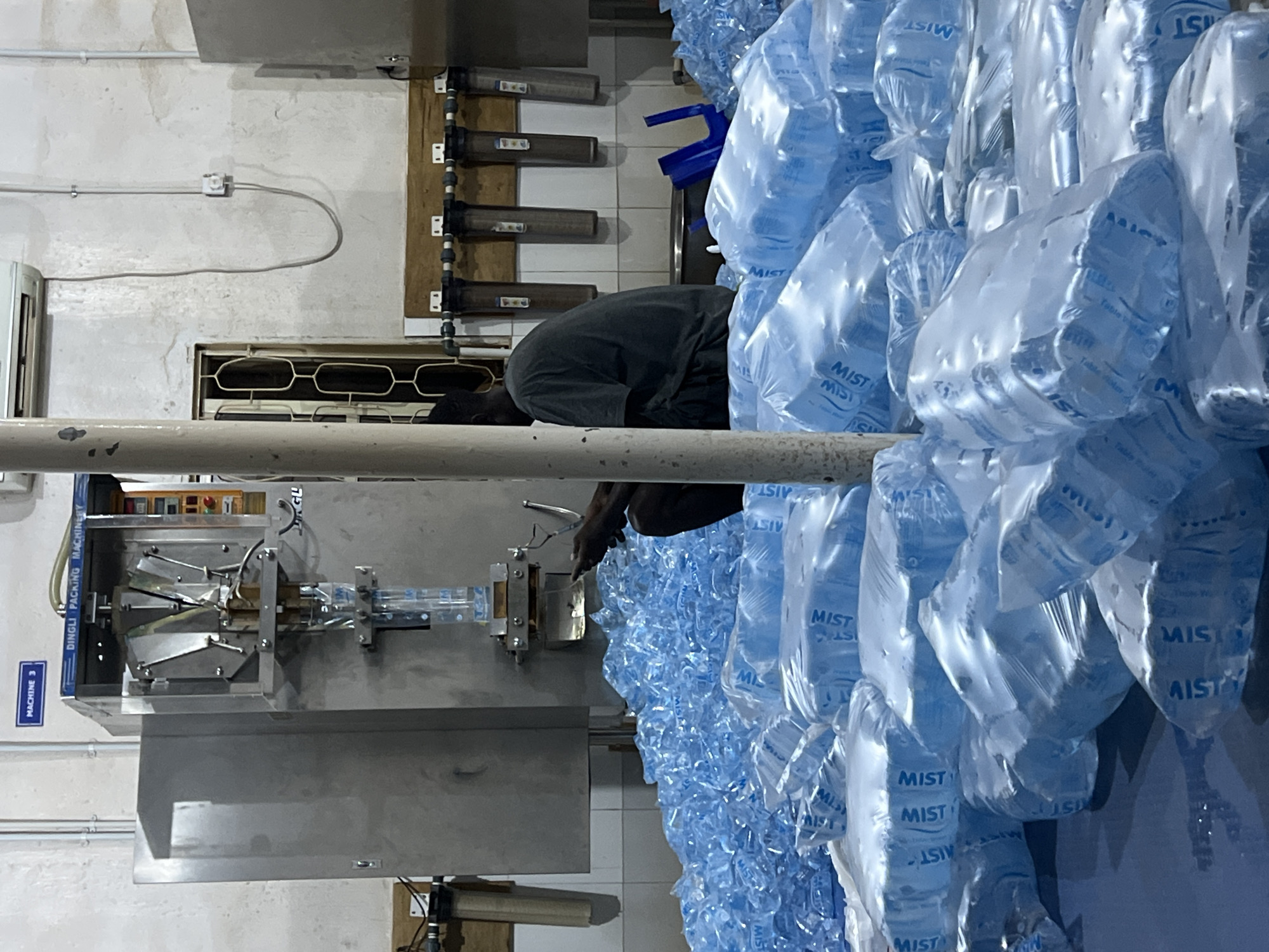 The image shows a man operating a Dingli packing machine (machine 3) in what appears to be a small factory setting, likely in Jos, Nigeria. He is handling and packing "MIST" brand water sachets. The background features a white wall, pipes, and possibly filtration systems. The environment appears clean and industrial. The focus is on the packing process of water sachets.