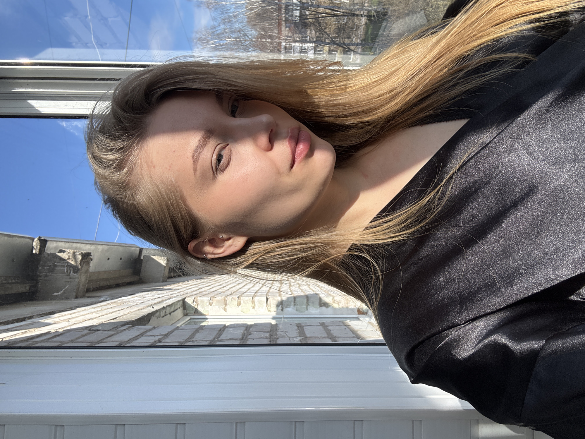 The image displays a close-up portrait of a young adult woman, presented at a 90-degree clockwise rotation. She looks directly at the camera with a calm, neutral expression, her head slightly tilted. Her long, light brown hair, with visible blonde highlights, frames her face and flows over her shoulders. She wears a black garment, possibly a top or dress, made of a shimmering, satin-like fabric that reflects the bright sunlight, creating highlights and shadows across its surface. A small, silver hoop earring is visible in her left ear.
The background, viewed through a window, shows a clear blue sky, indicating a sunny day. Bare tree branches are visible in the upper portion of the background, suggesting a time of year before leaves have fully bloomed, likely late winter or early spring. Beyond the branches and a distant horizon, indistinct building structures can be seen. Closer to the window, weathered concrete and brick elements, possibly part of an older building's roof or exterior, are visible. The strong, natural sunlight creates defined shadows on her face and hair. The overall setting appears to be indoors, near a window, looking out onto an urban or residential area, consistent with locations such as Chaikovsky, Russia. There is no visible text in the image.