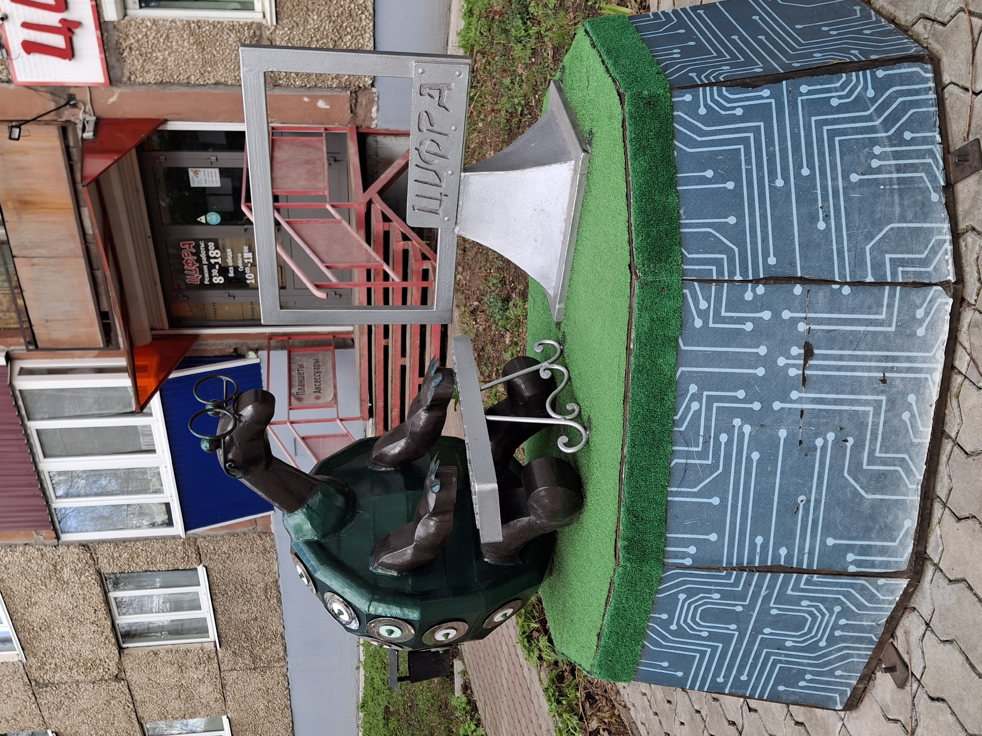 An outdoor photograph, taken in Sarapul, Russia, features a creative metal sculpture positioned on a sidewalk. The main subject is a stylized, dark green turtle wearing glasses, depicted operating a computer keyboard with its front limbs while looking at a monitor. The monitor's frame proudly displays the Russian word "ЦИФРА" (TSIFRA), which translates to "Digit" or "Number," in a decorative metallic font. The turtle's shell is adorned with several reflective, circular elements, adding a distinctive detail.
The sculpture rests on a patch of green, artificial turf, which is set upon a larger, greyish-blue base. This base is uniquely decorated with white circuit board patterns, emphasizing a theme of technology or computing.
In the background, a building with textured, light brown walls and multiple windows is visible. A prominent red-framed entrance belongs to a shop. A sign on the building's facade reiterates the name "ЦИФРА" at the top. Below this, the operating hours are listed as "Режим работы: 8:30-18:00 Без обеда" (Working hours: 8:30 AM - 6:00 PM, No lunch break), and for "Суббота: 10:00-14:00" (Saturday: 10:00 AM - 2:00 PM). A smaller sign indicates that the shop sells "Планшеты" (Tablets) and "Аксессуары" (Accessories). The scene appears to be set during daytime under an overcast sky, with pavement and sparse vegetation completing the outdoor setting.