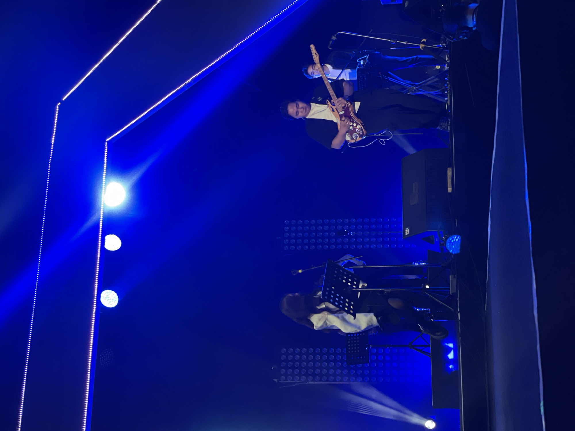 The image shows a concert stage with blue lighting. A man in a white shirt and black jacket is playing an electric guitar. Another person is behind him, partially obscured. Below them, a person wearing a white top and black pants is seated, possibly playing a keyboard or piano, with a music stand in front. The backdrop features LED lighting panels. Salembaran, Indonesia is not discernible from the provided image.