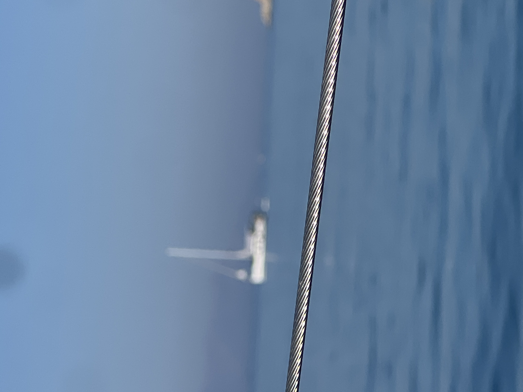 An out-of-focus image shows a sailboat on a calm blue sea under a hazy blue sky. In the foreground, a thick, metallic cable runs horizontally across the frame, also out of focus. The sailboat is distant and appears white with a tall mast and rigging. The cable is silver and shows a spiral texture formed by twisted wires. The overall impression is one of a vast, open body of water with a hint of human presence in the form of the sailboat and the foreground cable, which may be part of a boat's rigging or a dock. The location is listed as Unknown, Unknown.
