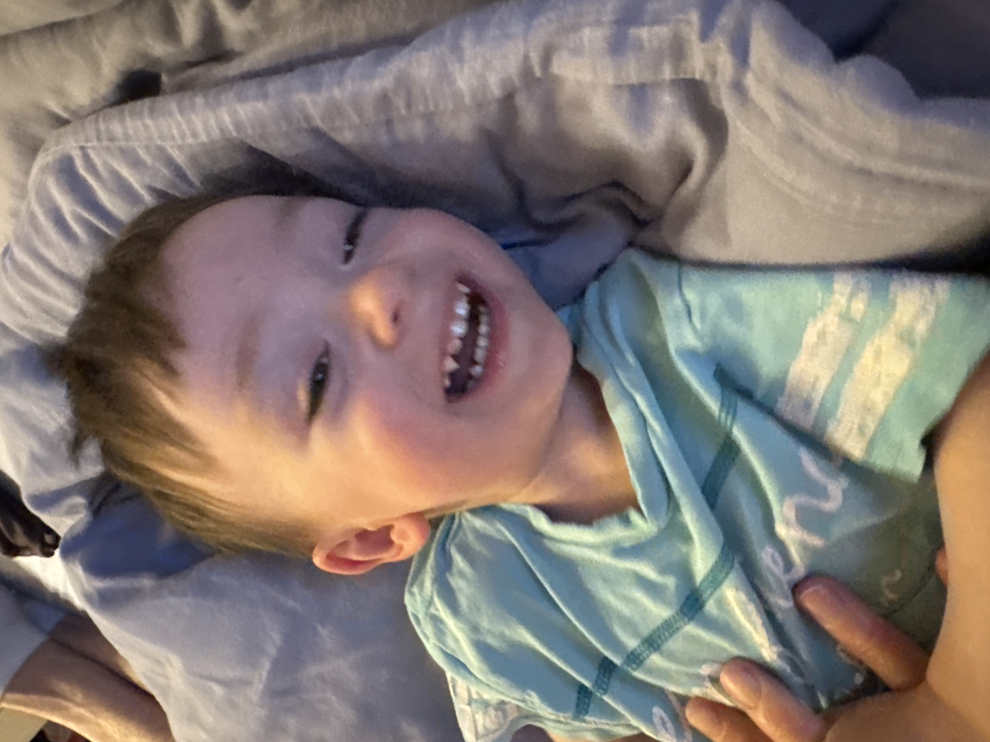 A young child is lying on their back on a bed or a cushioned surface, smiling widely and looking directly at the camera. The child has short brown hair, fair skin, and is wearing a light blue t-shirt with white and darker blue patterns. The background consists of light grey bedding. There is no indication of Sochi, Russia or any city setting.