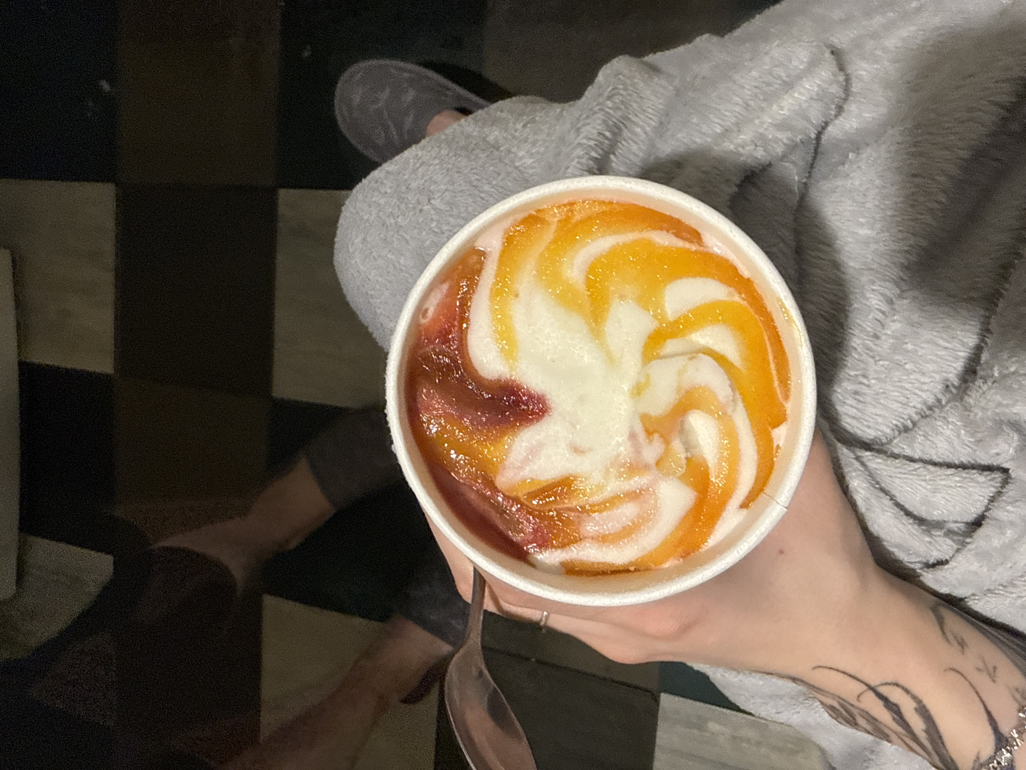 A person sits indoors, on a black and tan checkered floor. The person is wearing gray pants and a gray robe, as well as a gray slipper. They hold a cup of yogurt with a spoon in their right hand. The yogurt is swirled with red and orange syrup. The person has a tattoo on their right arm.
There is no evidence in the image to suggest the city of Moscow, Russia.