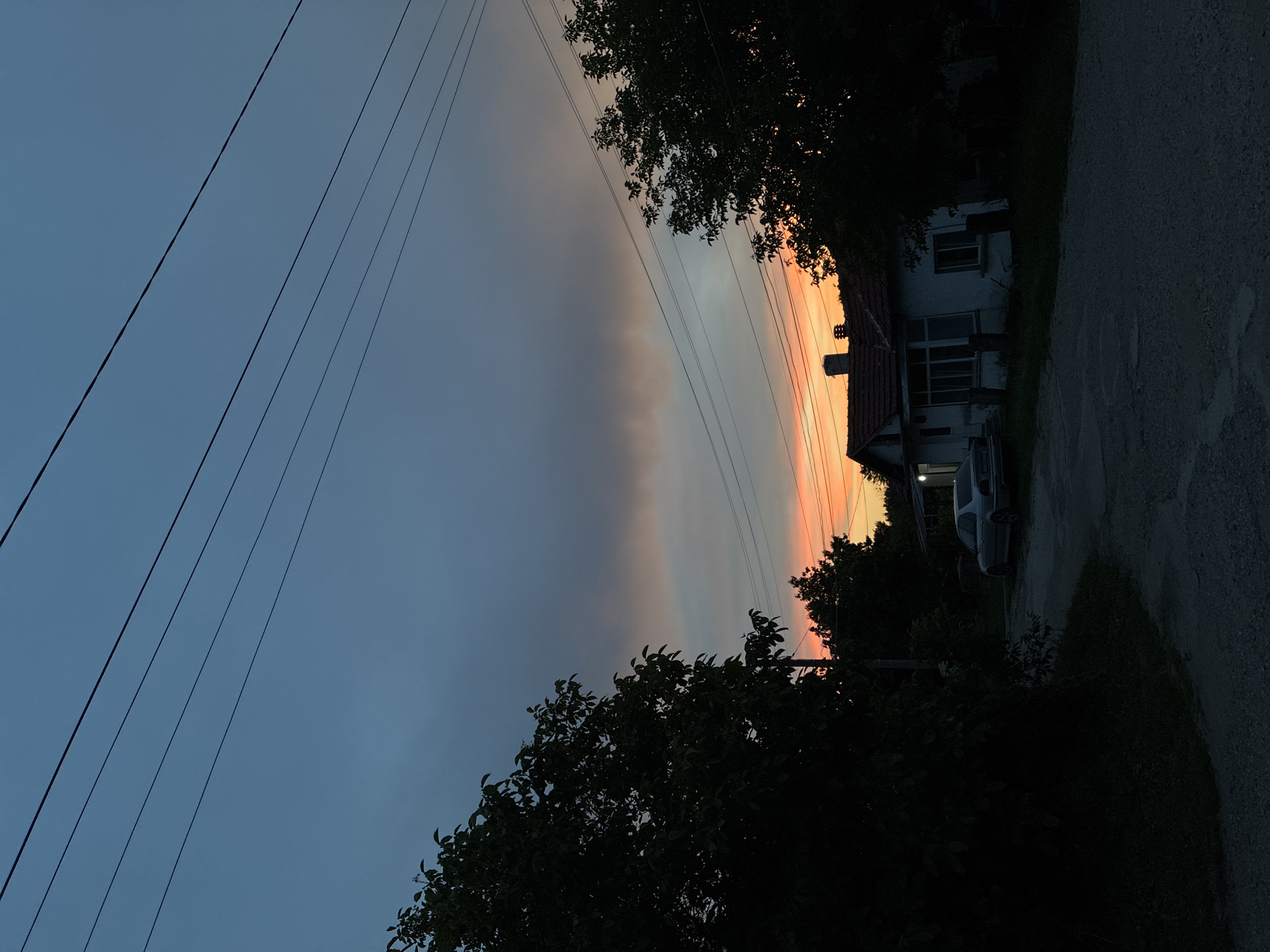 The image depicts a twilight scene in a rural area. The sky displays a gradient of colors, transitioning from a deep blue at the top to vibrant oranges and pinks near the horizon, indicating a sunset or sunrise. Silhouetted against this sky are power lines stretching across the frame. In the foreground, a paved road leads towards a modest house with a red-tiled roof. A white car is parked in front of the house. Dark trees and foliage frame the scene on both the left and right sides, adding to the atmospheric mood. The overall impression is one of quiet evening in a residential area.