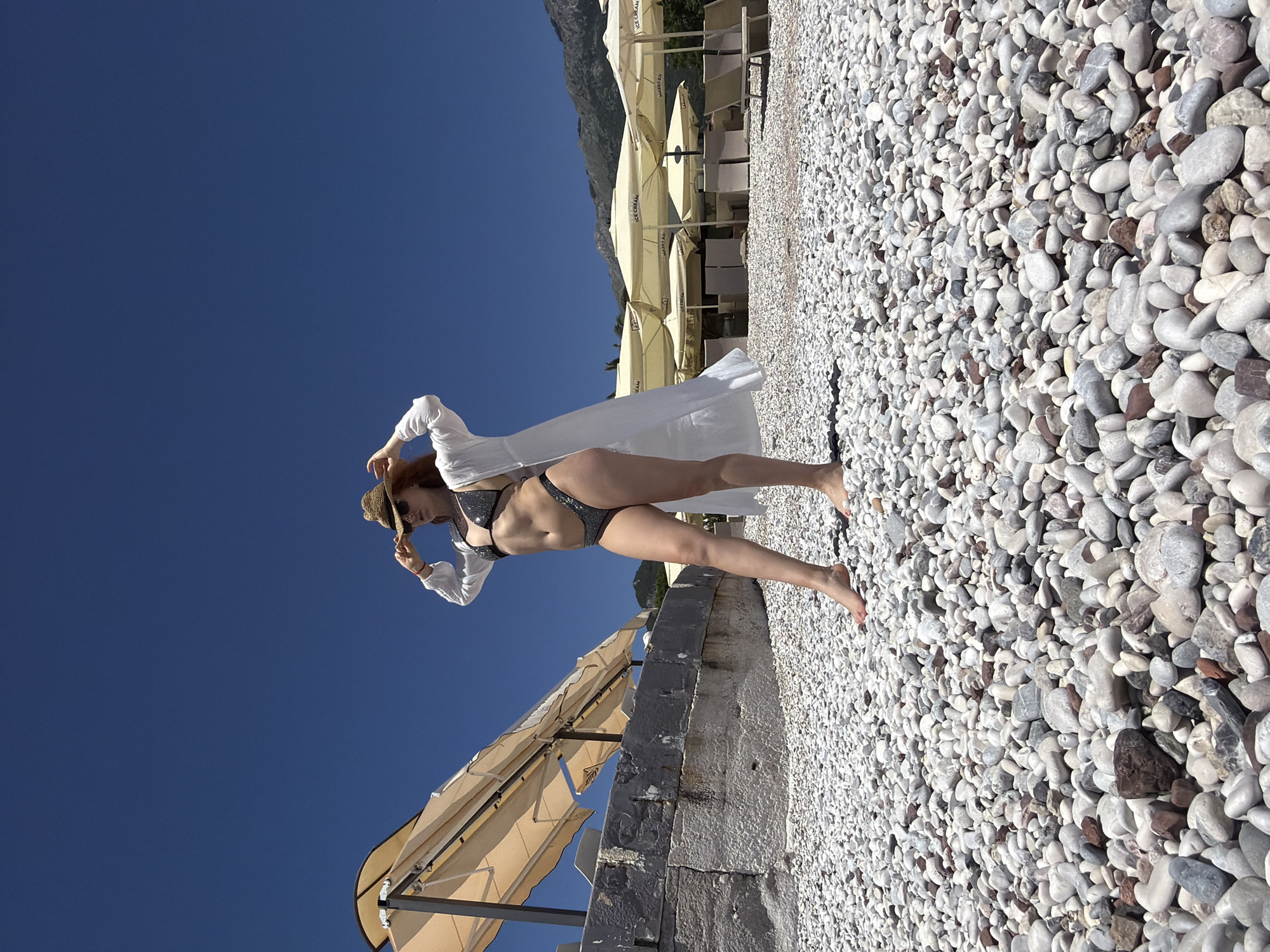 A woman wearing a dark bikini, a long white shirt, and a straw hat is standing on a rocky beach. She is holding the hat with one hand. The background consists of clear blue sky and beach umbrellas. The beach is made up of small, white rocks. The location is likely in Budva, Montenegro, given the coastal setting and clear blue sky.