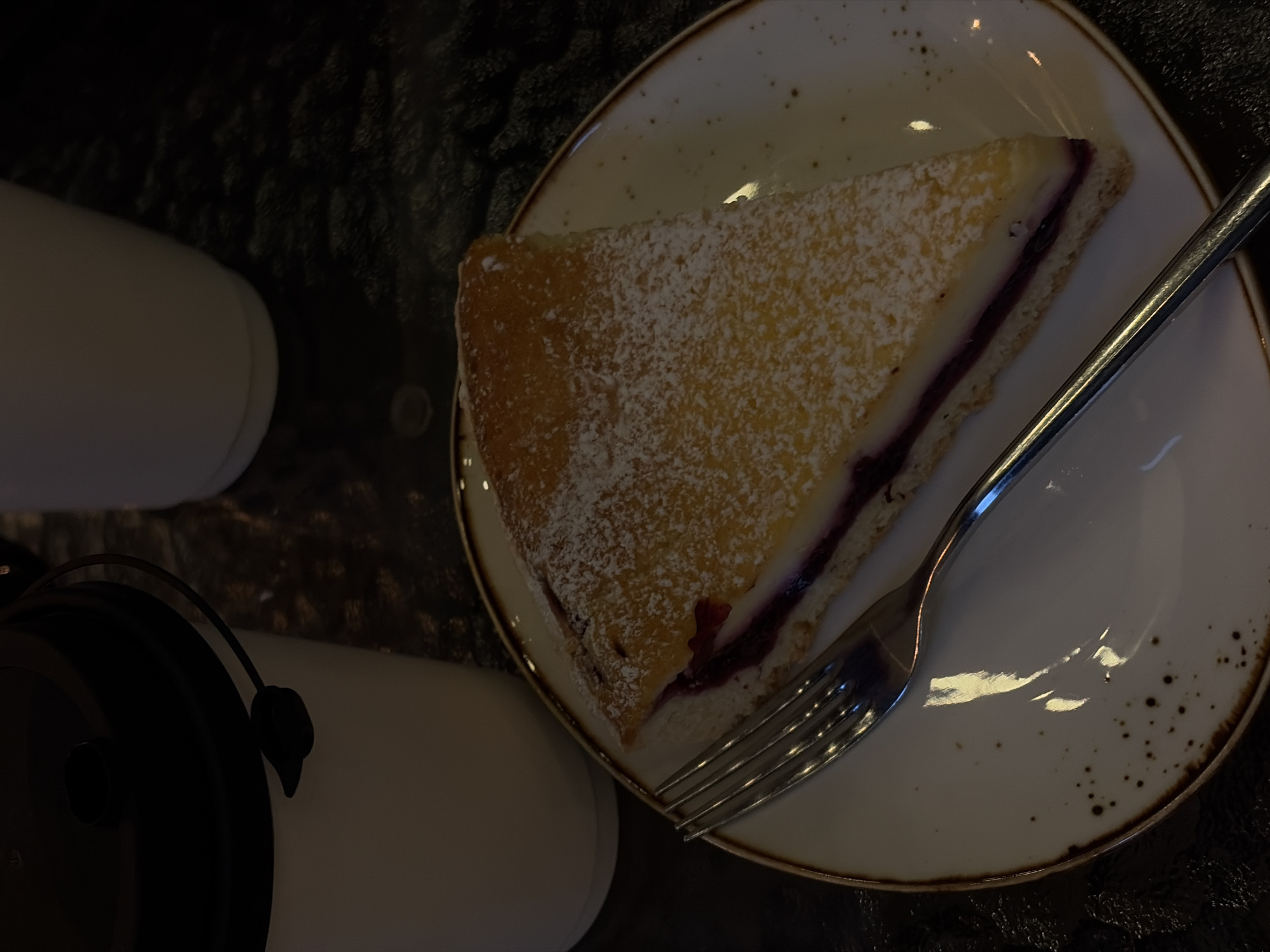 A close-up, overhead shot shows a slice of cake on a plate with a fork, accompanied by two cups, against a dark, textured background. The cake slice is triangular, dusted with powdered sugar, and has a layer of what appears to be a dark fruit filling. A silver fork rests on the right side of the plate, partially covering the cake. The plate is white with a speckled brown rim. To the left, there are two white cups; one is taller and cylindrical, while the other is shorter and wider with a black lid and a curved handle. The background appears to be a dark, possibly rough surface. There's no indication of the location being Novopokrovskaya, Russia within the frame. No people are visible, and there are no actions or interactions occurring within the scene.