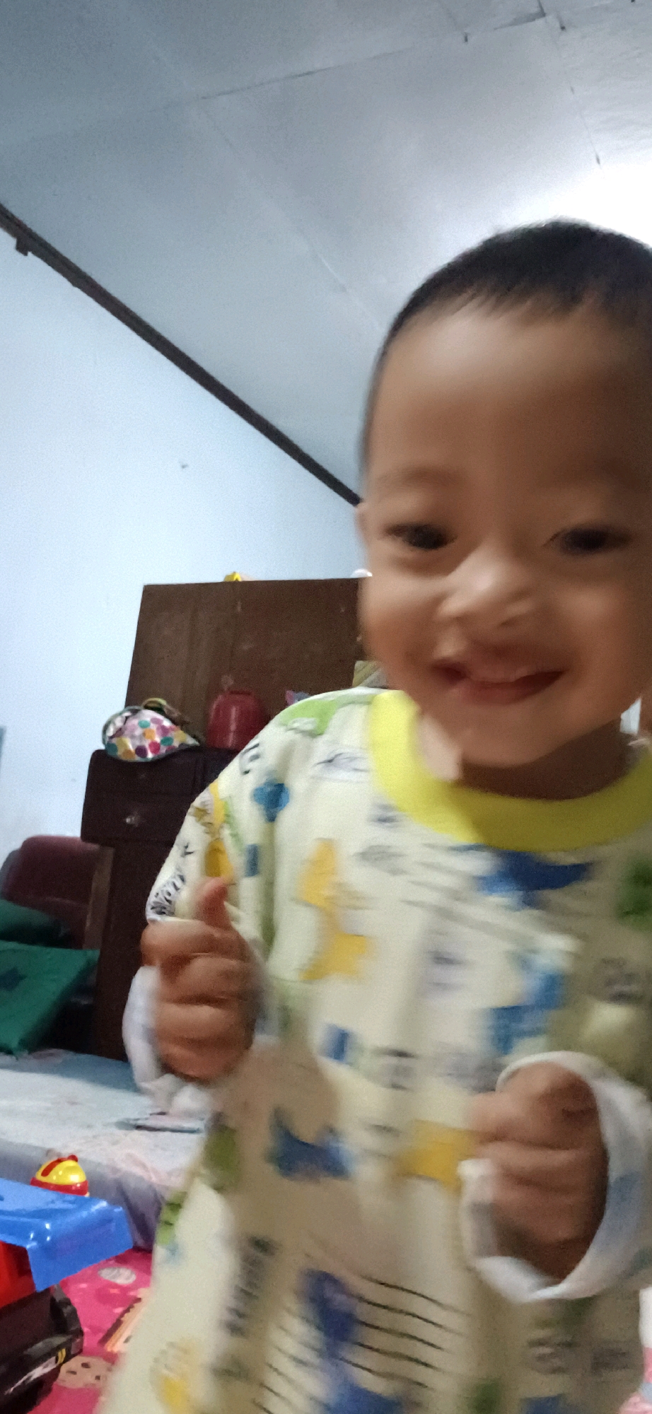 The image depicts a young child, likely a toddler, with dark hair and brown eyes, smiling broadly at the camera. The child is wearing a long-sleeved shirt with a yellow collar and a light-colored pattern featuring blue and yellow shapes that appear to be animals or cartoon characters. The child's right hand is raised, with the thumb extended upwards in a gesture that could be interpreted as a "thumbs up."
The child is positioned in what appears to be an indoor setting. Behind the child, a dark wooden cabinet or dresser is visible. On top of this furniture, a multi-colored, possibly fabric, item with polka dots is placed, along with a red cylindrical object. A wall, painted a light blue, forms the background. To the left and foreground, portions of what seem to be children's toys are present, including a red and yellow toy with a dome shape and a blue plastic component. The floor appears to be covered with a patterned fabric or rug, and there are also green cushions visible. The overall lighting suggests an indoor environment, possibly during the day. The image quality is somewhat blurred, indicating it may have been taken with a mobile device or under low light conditions.
This scene is located within a residential interior in the city of Wonokerso, Indonesia.