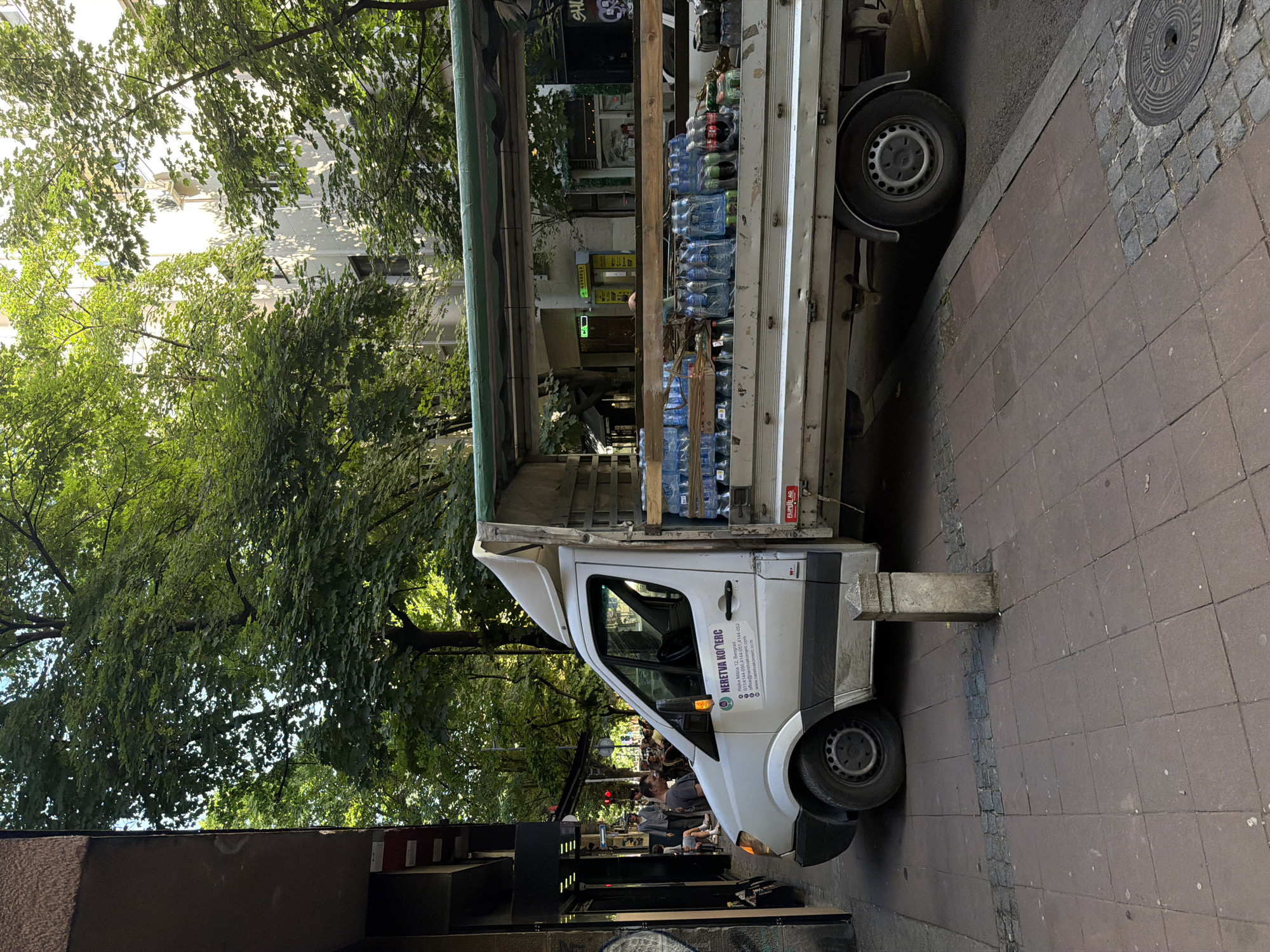 The image depicts a white delivery truck parked on a sidewalk in Belgrade, Serbia. The truck's cargo bed is stacked with bottled water, and it appears to be stopped in front of a commercial establishment, possibly delivering goods. Text on the truck identifies "NERETVA KOMERC," a company in Belgrade, along with contact information. The surrounding environment includes lush green trees, indicating a tree-lined street. People are visible near the cafe. The sidewalk is paved with tiles and edged with a curb.