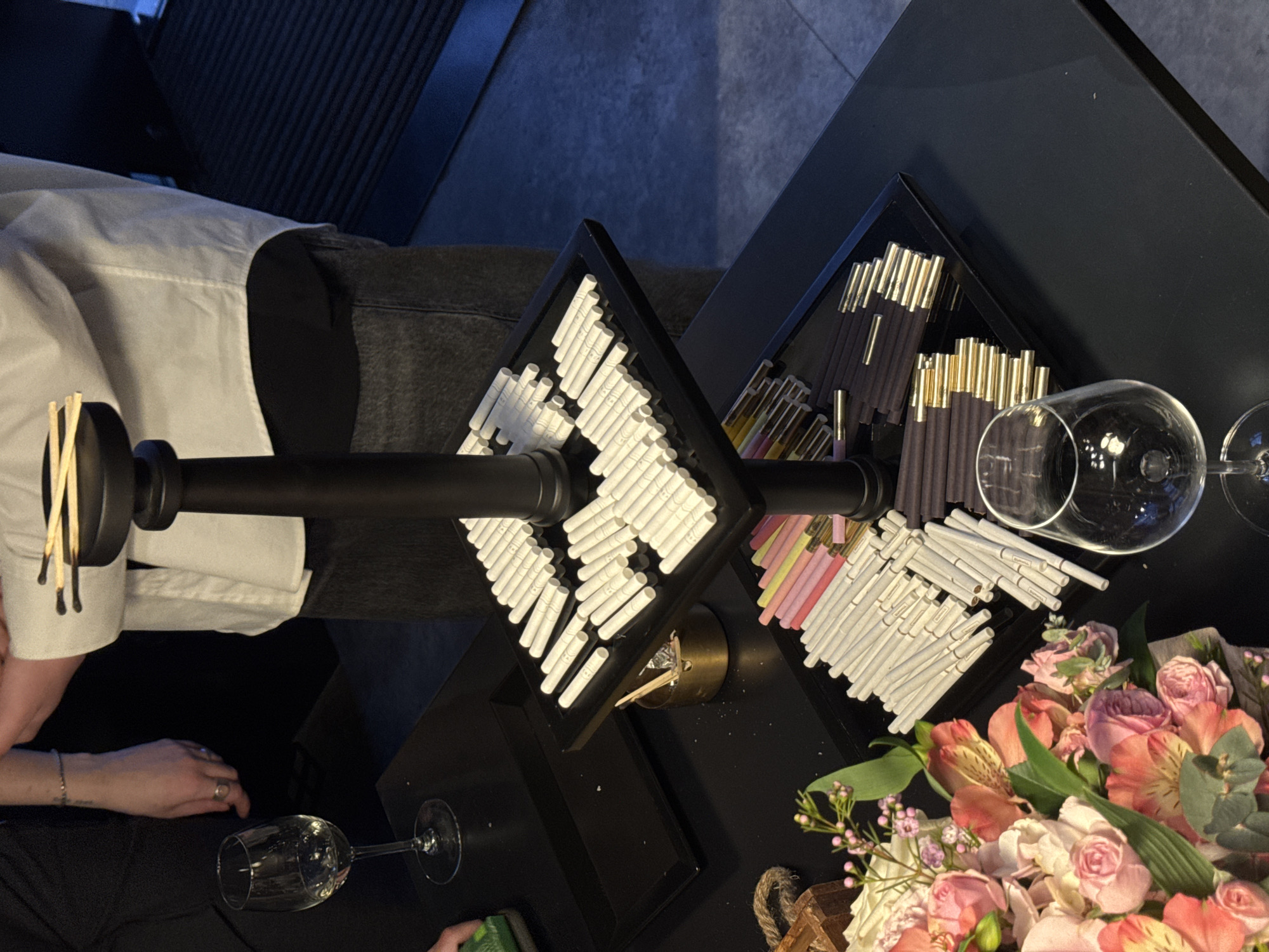 An indoor scene depicts an event or retail display. Two individuals are partially visible. On the right, a person wearing a white collared shirt over a dark undershirt and dark trousers is positioned behind a black three-tiered display stand. This person's right hand is visible at the top tier, holding several light-colored wooden matches. On the left, another person's left hand, adorned with a ring and a bracelet, holds a small green object near a clear wine glass containing a clear liquid.
The central object is a black, three-tiered display stand. Its top tier holds several light-colored wooden matchsticks. The middle tier contains multiple rows of white, slender cylindrical objects, consistent with nicotine or heated tobacco sticks. The bottom tier features additional rows of similar cylindrical products: one section of white sticks, another section of pastel-colored (pink, yellow, green) sticks with gold tips, and a third section of dark brown/black sticks with gold tips.
On the black table surface, to the left of the stand's middle tier, is a small, brass-toned cylindrical container functioning as an ashtray, containing used matchsticks. A dark rectangular tray is positioned further left. In the lower left foreground, a vibrant bouquet of fresh flowers, including pink and peach roses and alstroemeria, is present. Another empty clear wine glass with a stem rests on the table in the lower right foreground. The immediate environment is characterized by dark furnishings, including the black table and display unit, under ambient lighting. A dark, ribbed mat or floor covering is visible in the background to the right. The image does not contain specific visual identifiers to pinpoint a precise location within Sofia, Bulgaria.