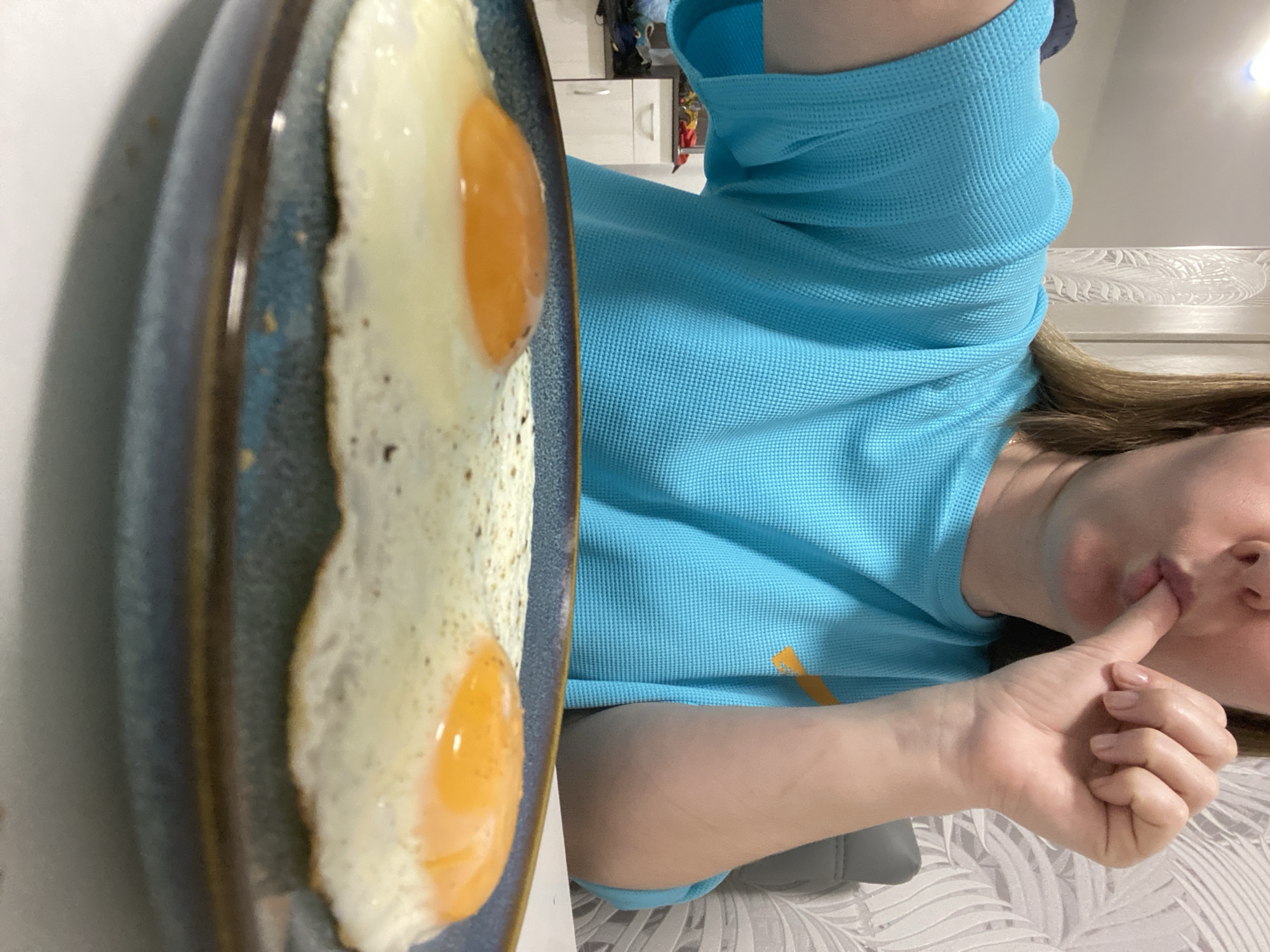 A woman in a light blue shirt is seen with a plate of two fried eggs next to her. She is holding a finger to her lips. The background includes a white wall and a patterned surface, possibly a bed or couch. Some kitchen elements are visible in the distance. No information about the location in the city of Noviy, Russia is available.