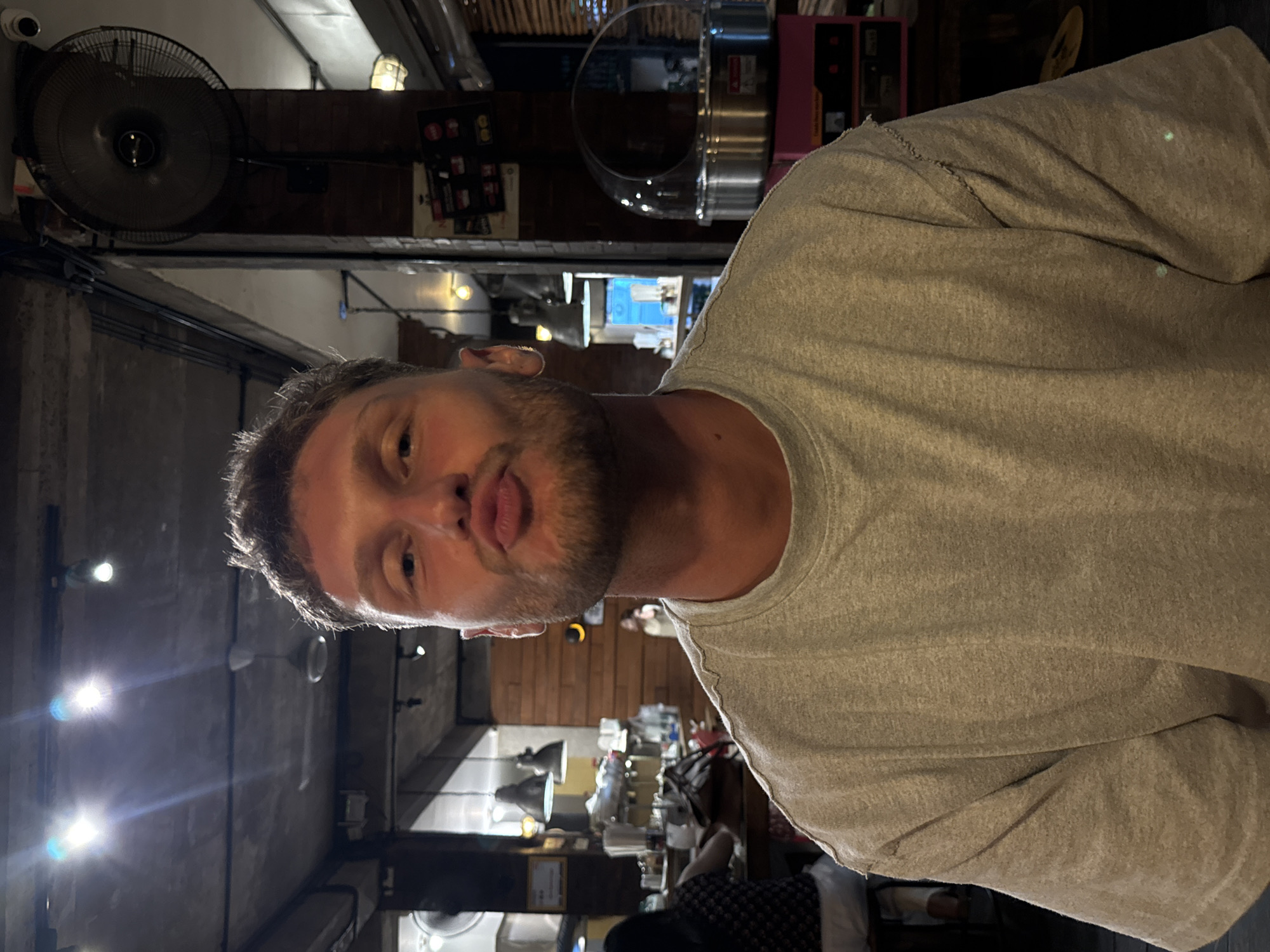 A man with short, light-colored hair and a beard is indoors, wearing a gray t-shirt. He is positioned at a slight angle, with the left side of his face closer to the camera. He is making a kissing face, and his eyes are light in color.
Behind him, the setting appears to be a restaurant or cafe. There are tables and chairs, and other people are present in the background, though they are out of focus. There is an industrial-style fan mounted on the ceiling, and various pieces of equipment and signage can be seen on the walls. The lighting is somewhat dim, with bright spots that could be from light fixtures.
Based on the details visible in the background, this is likely taken in North Kuta, Indonesia, given the local style.