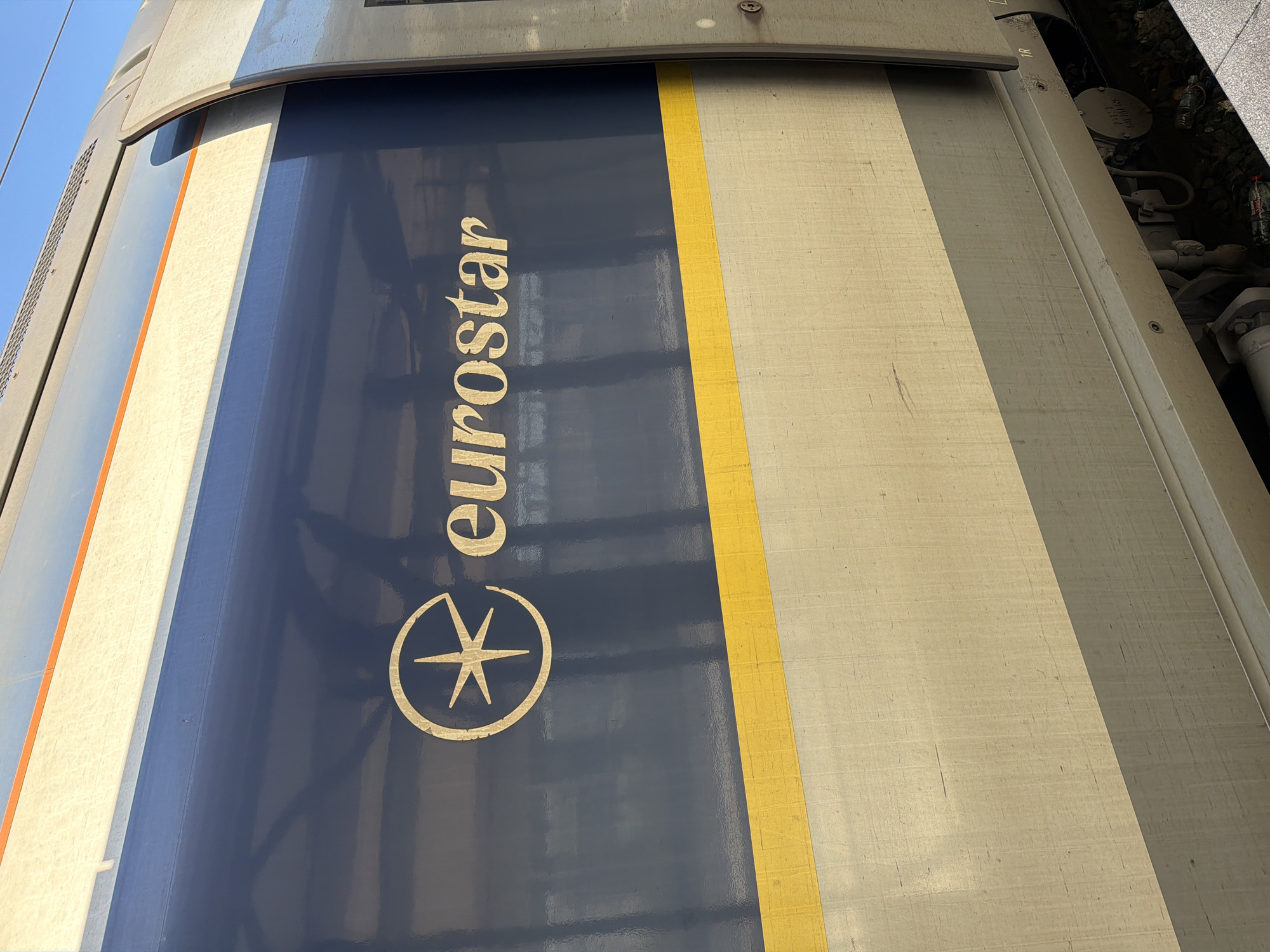 This close-up shot depicts the side of a Eurostar train. The Eurostar logo, featuring the word "eurostar" in gold lettering above a stylized compass rose, is prominently displayed on a dark blue background. A yellow stripe runs vertically alongside the blue section. The train's body appears to be a textured grey-beige. Visible in the background are parts of the train's undercarriage. The specific location within Paris is not discernable from the image.