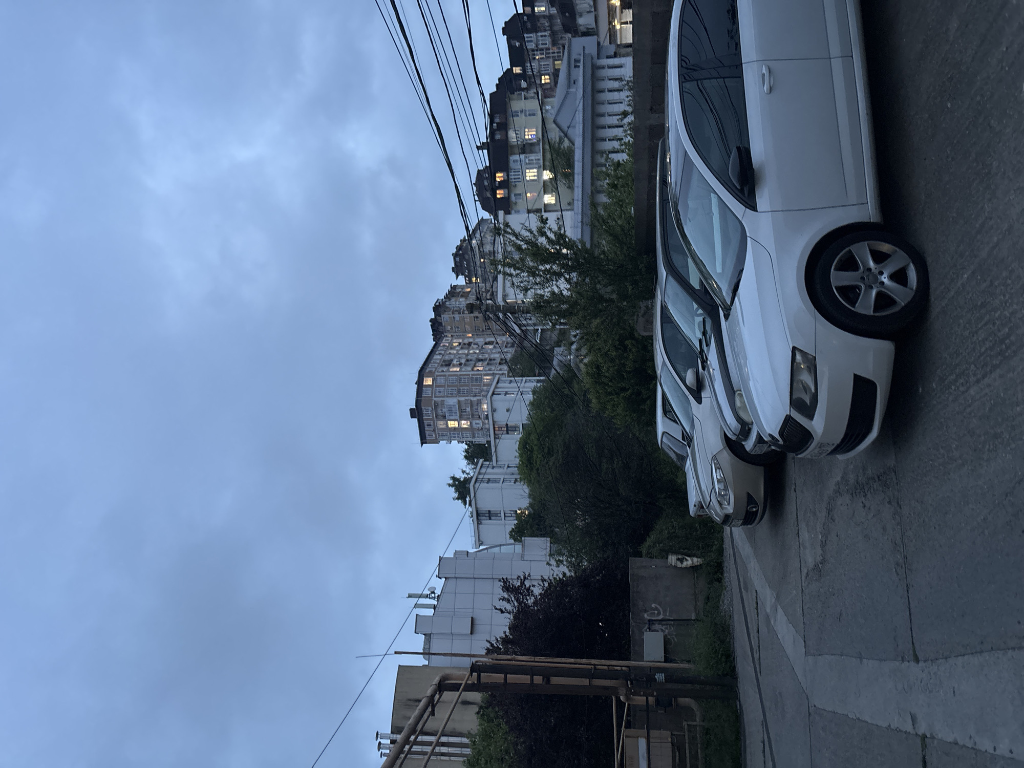 The image shows a street scene in what appears to be the city of Sochi, Russia, based on architectural characteristics. Several multi-story residential buildings, some with illuminated windows, are visible in the background. A row of parked white cars occupies the right side of the frame, adjacent to a curb. Overhead, power lines stretch across the sky. The overall light suggests it is either dusk or dawn, under an overcast sky. Near the bottom of the image, a section of industrial piping is visible, indicating a mixed residential and industrial or utility zone. No people are visible in the image.