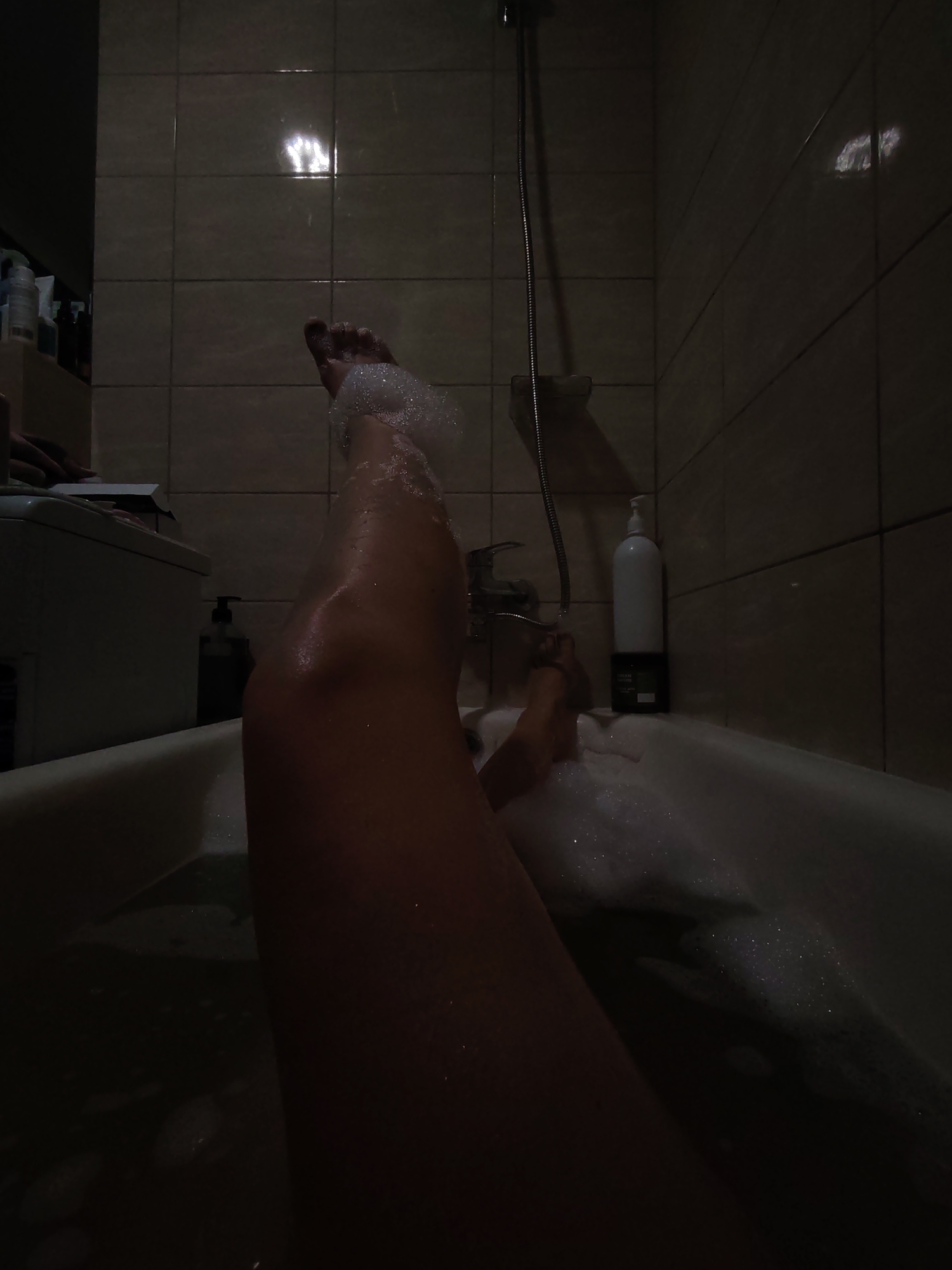 The image depicts a person's legs and feet submerged in a bathtub filled with foamy water. The setting appears to be a bathroom with tiled walls and a showerhead visible. There are bottles of toiletries on a shelf to the left and a large white pump bottle of body wash on the right. The lighting is dim, creating shadows and highlighting the water and bubbles. There is no indication of the location being in Izhevsk, Russia.