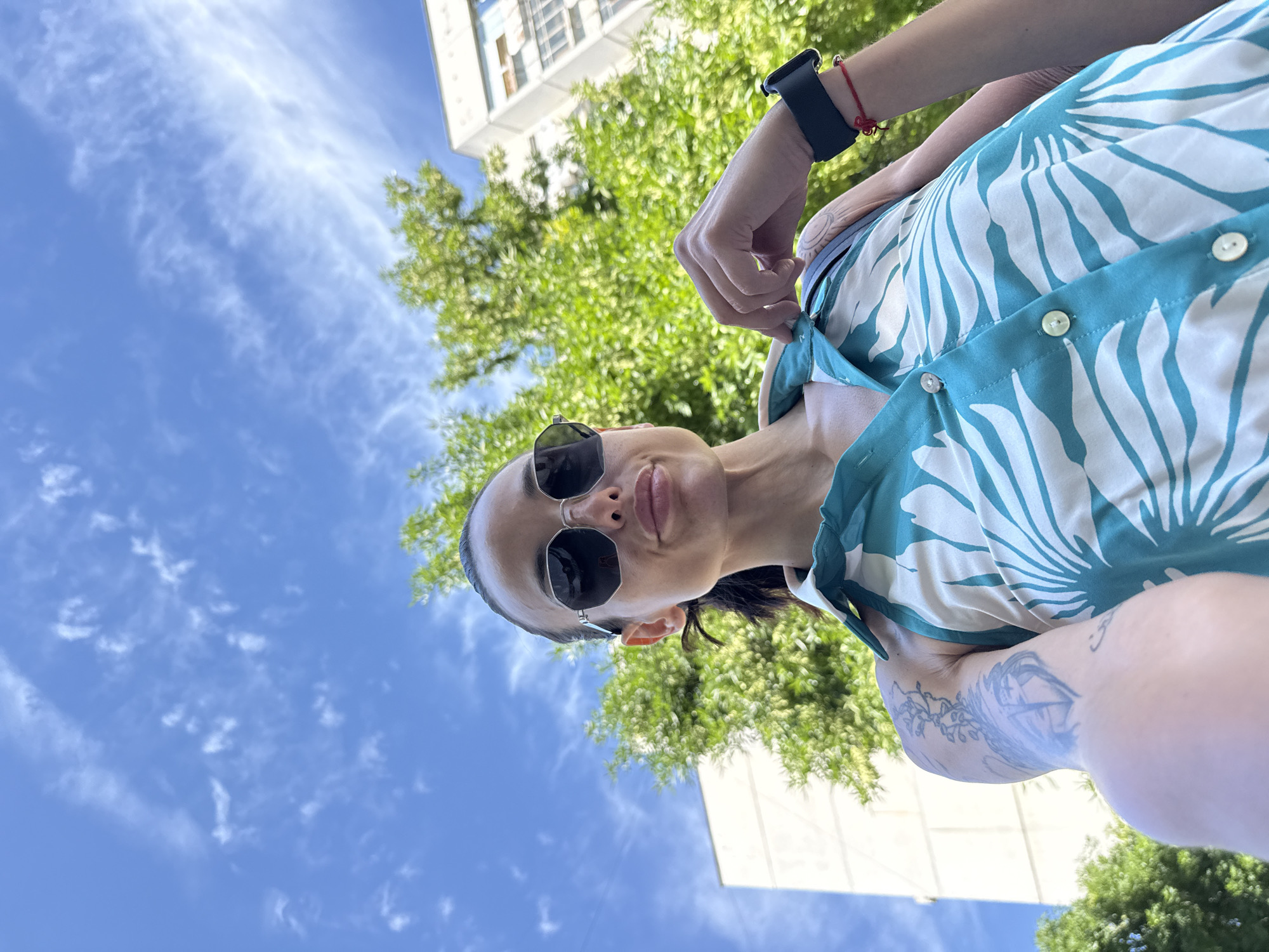 The media file captures a low-angle, close-up, selfie-style shot of a person wearing sunglasses and a teal and white patterned shirt. The person is looking up towards the camera, with their lips slightly parted. They have a dark smartwatch on their left wrist, adorned with a red string bracelet. The background consists of a bright blue sky with wispy white clouds, lush green trees, and the corner of a white building. The lighting suggests it is daytime, likely sunny. The overall mood appears relaxed and casual. There are tattoos visible on the person's left arm. The scene appears to be set outdoors in an urban or suburban environment.