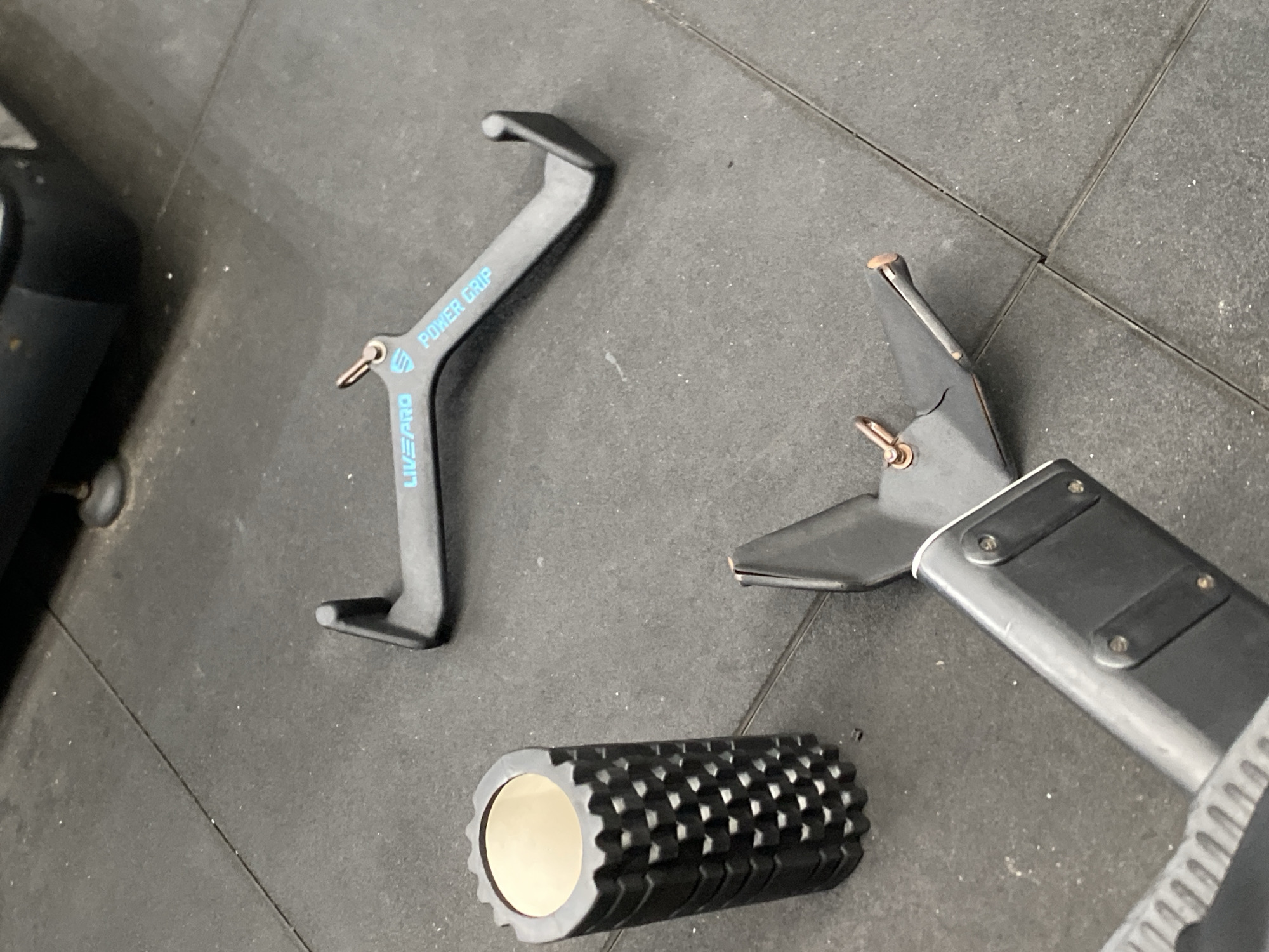 The image shows gym equipment on a black rubber floor. In the foreground, a portion of a weight machine is visible. To the left, a black foam roller with a textured surface is present. In the middle ground, there are two black gym attachments. One is a V-shaped handle with blue text that reads "LIVEPRO POWER GRIP." The other is a U-shaped attachment with a pin. The background shows more of the gym floor and possibly another piece of equipment. The lighting suggests an indoor setting, likely a gym.