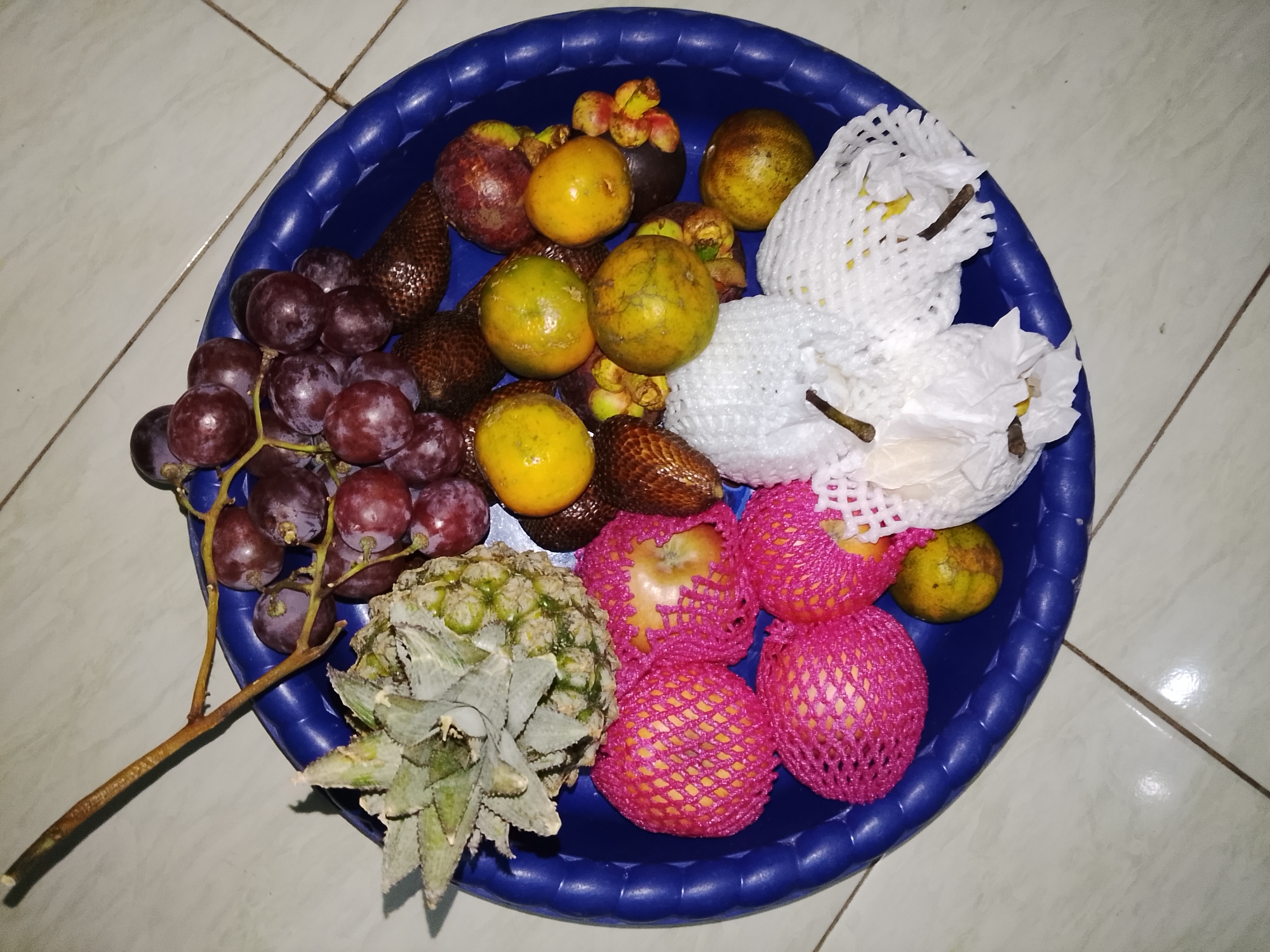 A close-up, overhead shot captures a collection of assorted fruits arranged in a blue plastic basin. The fruits include a bunch of dark red grapes, several small, yellow-orange fruits, some mangosteens, a pineapple, and several other fruits wrapped in white and pink protective netting. The basin is placed on a light-colored tiled surface. The scene appears to be indoors, possibly in a home. The image is likely from Dawuan, Indonesia, based on the context provided. No people or animals are visible in the image.