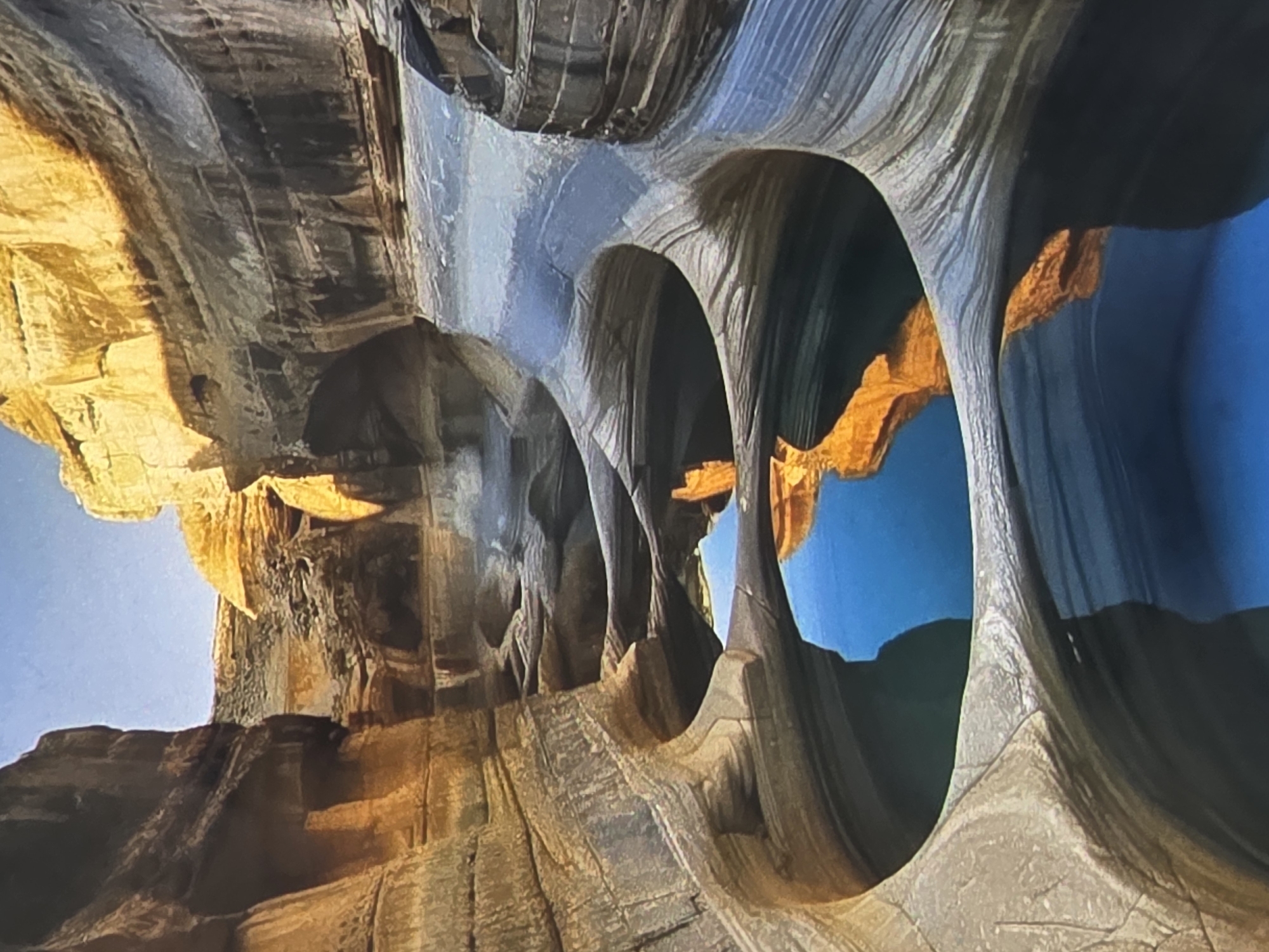 The image displays a natural rock formation with water pools. The rocks are layered and appear eroded, with smooth, curved surfaces. Sunlight illuminates parts of the rock, creating highlights and shadows. Several interconnected pools of clear blue water are visible within the rock formations, reflecting the sky and the rock walls. The scene suggests a natural landscape, possibly a canyon or a gorge, with water carving through the stone over time. There are no buildings, structures, or any indications of urban environment. Therefore, it is not possible to associate this image with the city of Kyiv, Ukraine.