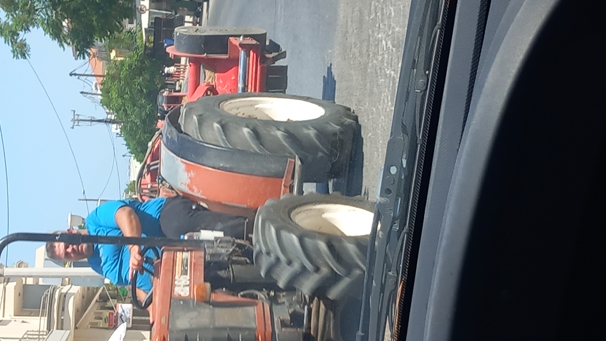 A man is driving a red tractor on a street in Kos, Greece. The man is wearing a blue t-shirt and black pants. The tractor is pulling a red piece of farm equipment. The street is paved and there are buildings on the side of the road. The sky is blue and there are some clouds. There are also trees and utility poles in the background. The image is taken from inside a vehicle, looking out through the windshield.