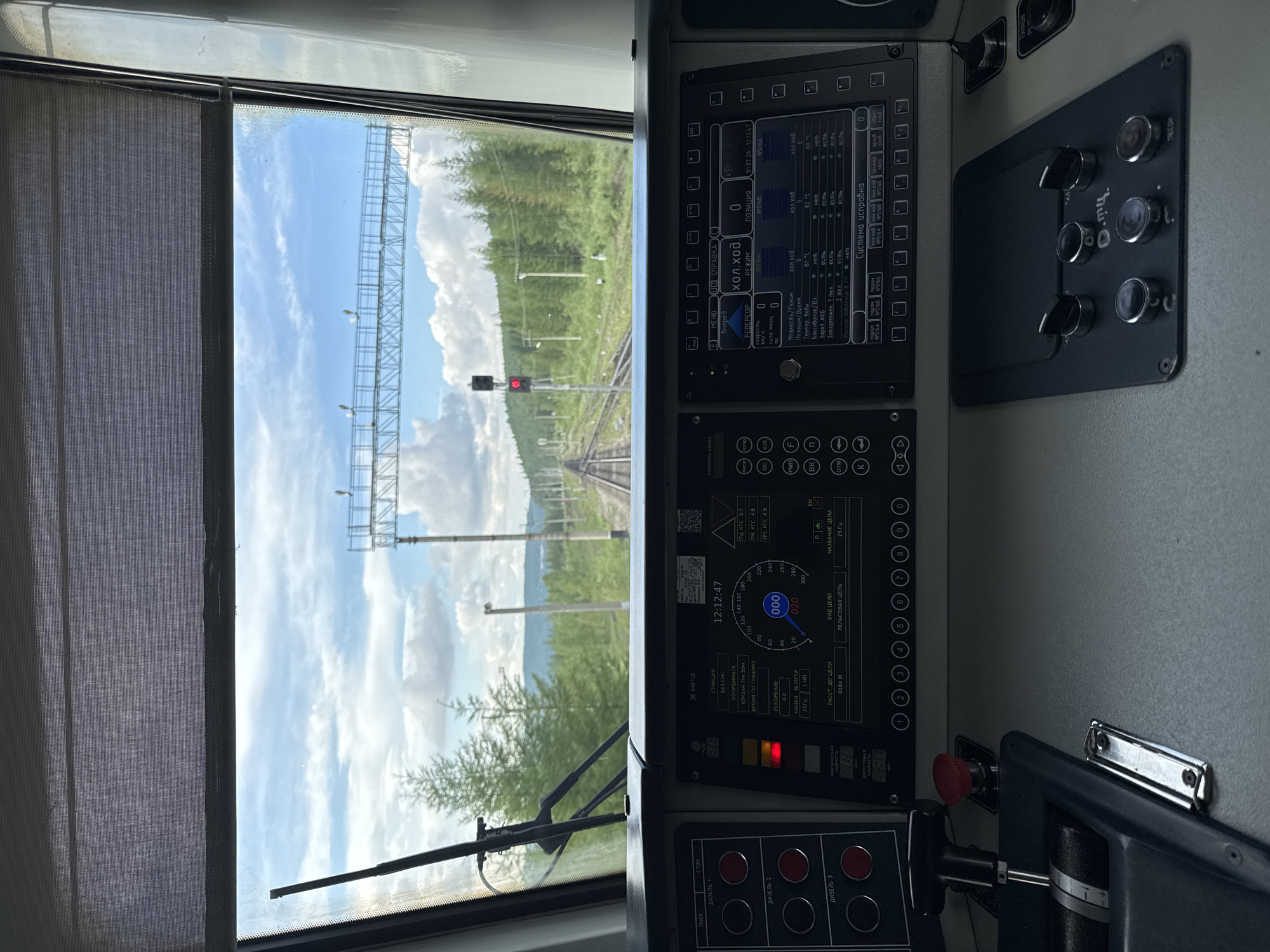 The image depicts the interior of a train cab, looking out through the windshield at a railway track stretching into the distance, surrounded by a forest under a cloudy sky. The foreground is dominated by the train's control panel, featuring various buttons, levers, and digital displays. On the left side of the control panel, there are several large circular buttons labeled "ПУСК" (Start), "СТОП" (Stop), and "ДИЗЕЛЬ 1", "ДИЗЕЛЬ 2", "ДИЗЕЛЬ 3". Below these are two circular buttons and a lever. A red emergency stop button is also visible. The central part of the control panel displays a digital screen showing train information. Key readings include "СТАНЦИЯ" (Station) as "БЕЗ СНС" (Without SNS), "КООРДИНАТА" (Coordinate) as "0341км 7лк 50м" (0341km 7th side 50m), "ВРЕМЯ ПО ГРАФИКУ" (Time according to schedule) as "0.0", and "УСКОРЕНИЕ" (Acceleration) as "0.0". It also shows "КАНАЛ № ПУТИ" (Track channel) as "25 Гц" (25 Hz) and "1 НП" (1 NP). A speed indicator dial shows a reading around 20 km/h, with the number "020" highlighted in a circle. Below this, "ФАКТИЧЕСКАЯ СКОРОСТЬ" (Actual speed) displays "0.00 km/h". Further displays read "РАССТ. ДО ЦЕЛИ" (Distance to destination) as "0184 M" (0184 meters), "ВИД ЦЕЛИ" (Type of destination) as "РЕЛЬСОВАЯ ЦЕПЬ" (Track circuit), and "НАЗВАНИЕ ЦЕЛИ" (Name of destination) as "25 Гц" (25 Hz). To the right, another digital display screen shows "ХОЛ. ХОД" (Idle mode) with multiple entries, including "Мощность/Режим" (Power/Mode) as "0", "Темпер. Воды" (Water temperature) as "81 °C", "Боксование/Бз" (Skidding/Bz) as "нет" (no), "Заторможен 1 мед" (Braked 1 second) as "2 мед" (2 seconds), and "Наклон/Бакет" (Incline/Bucket) as "5 Гр" (5 degrees). Other parameters like "Датчик" (Sensor), "ЭКСПЛУАТАЦИИ" (Operation), and "СИСТЕМА ИСПРАВНА" (System is OK) are also visible. The view through the windshield shows a double railway track disappearing into the horizon. The tracks are flanked by a dense forest of coniferous trees. In the distance, a signal light is visible, currently showing red. Overhead, a railway signal gantry with lights spans the tracks. The sky is partly cloudy with a bright, diffused light, suggesting daytime. The context provided places this scene in Zolotinka, Russia. The operational status and labeling of the control panel suggest this is a working train cab, likely in a remote or rural area given the surrounding forest. There are no people visible in the image. The overall impression is one of a functional and operational train environment, poised for
