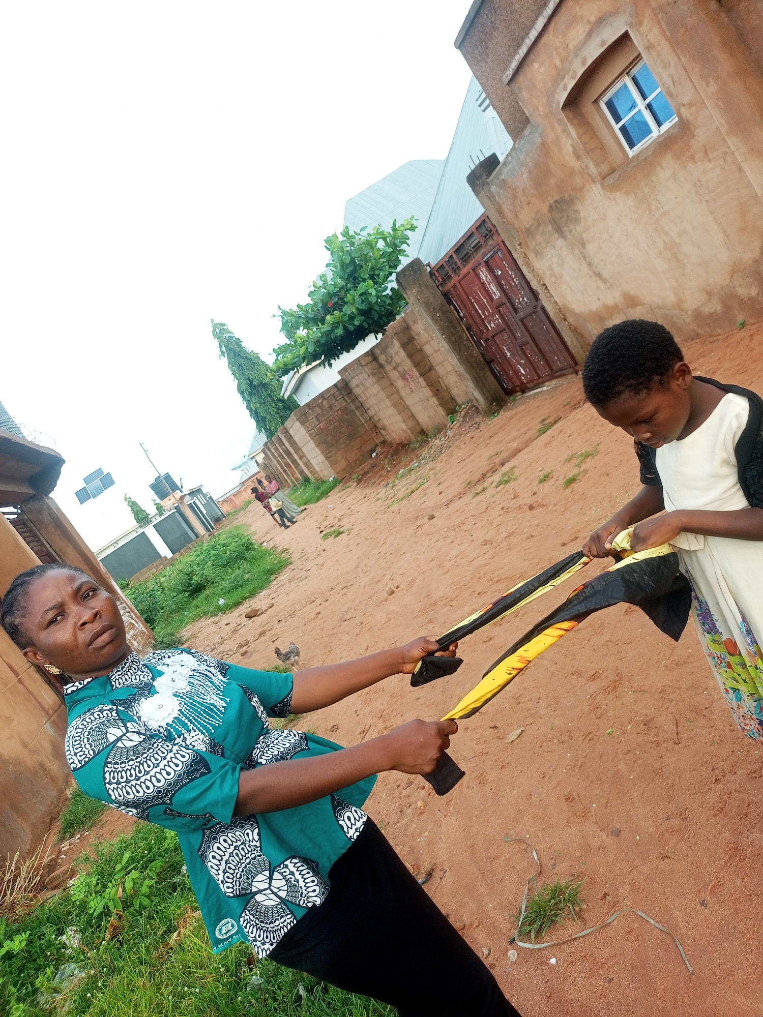 In Tempure, Nigeria, two individuals are engaged in an interaction in an outdoor environment characterized by a dusty, sandy ground. On the left, a woman with braided hair, wearing a green patterned blouse and black pants, holds a piece of fabric. To her right, a child, wearing a white dress with black detailing and a floral skirt, is also interacting with the same fabric. The fabric appears to be a long strip with alternating black and yellow sections. In the background, there are structures that suggest a residential area, including a tan-colored building with a blue-paned window, a weathered red door, and a brick wall. Green vegetation, including grass and trees, is visible on the left side and further in the background. The sky is overcast. A chicken is visible on the ground in the mid-ground.