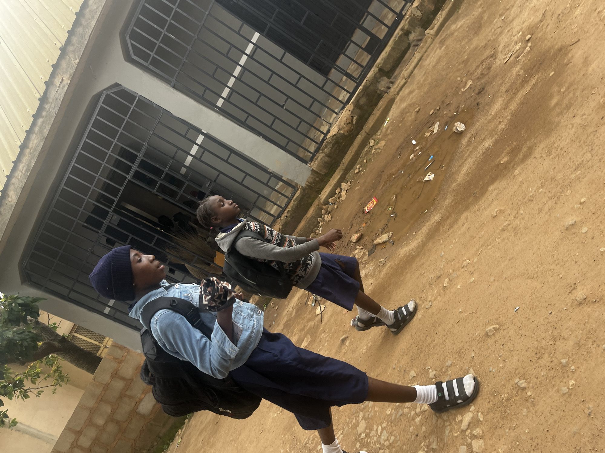 In Gura Top, Nigeria, two young girls walk along a dirt path. The girl in the foreground, on the left, wears a blue denim jacket, a navy blue beanie, and dark blue shorts with white socks and black sandals. She carries a large black backpack and a patterned cloth in her left hand. The girl behind her, on the right, wears a grey hooded sweatshirt with a patterned design and dark blue shorts, also with white socks and black sandals. She carries a black backpack. They are walking past a building with barred windows and a metal roof. The ground is uneven, with scattered debris and puddles of water.