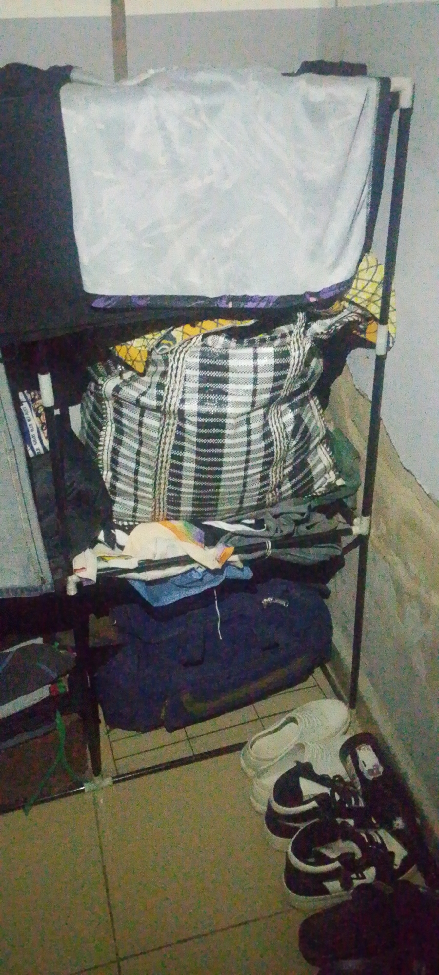The image shows a corner of a room with a black metal clothing rack filled with various items. The top shelf has a large white tarp or sheet draped over it. Below that, a black, white, and grey checkered duffel bag is prominent, overflowing with what appears to be fabric. Underneath the duffel bag are piles of folded clothing in dark colors, including what looks like a navy blue jacket or sweater. On the floor, a pair of white slip-on shoes is visible, next to a pair of black and white sneakers. The room's wall is a light, textured gray, and the floor appears to be tiled. The lighting is dim. This scene is likely located within a residence in Jauro Yinu, Nigeria.
