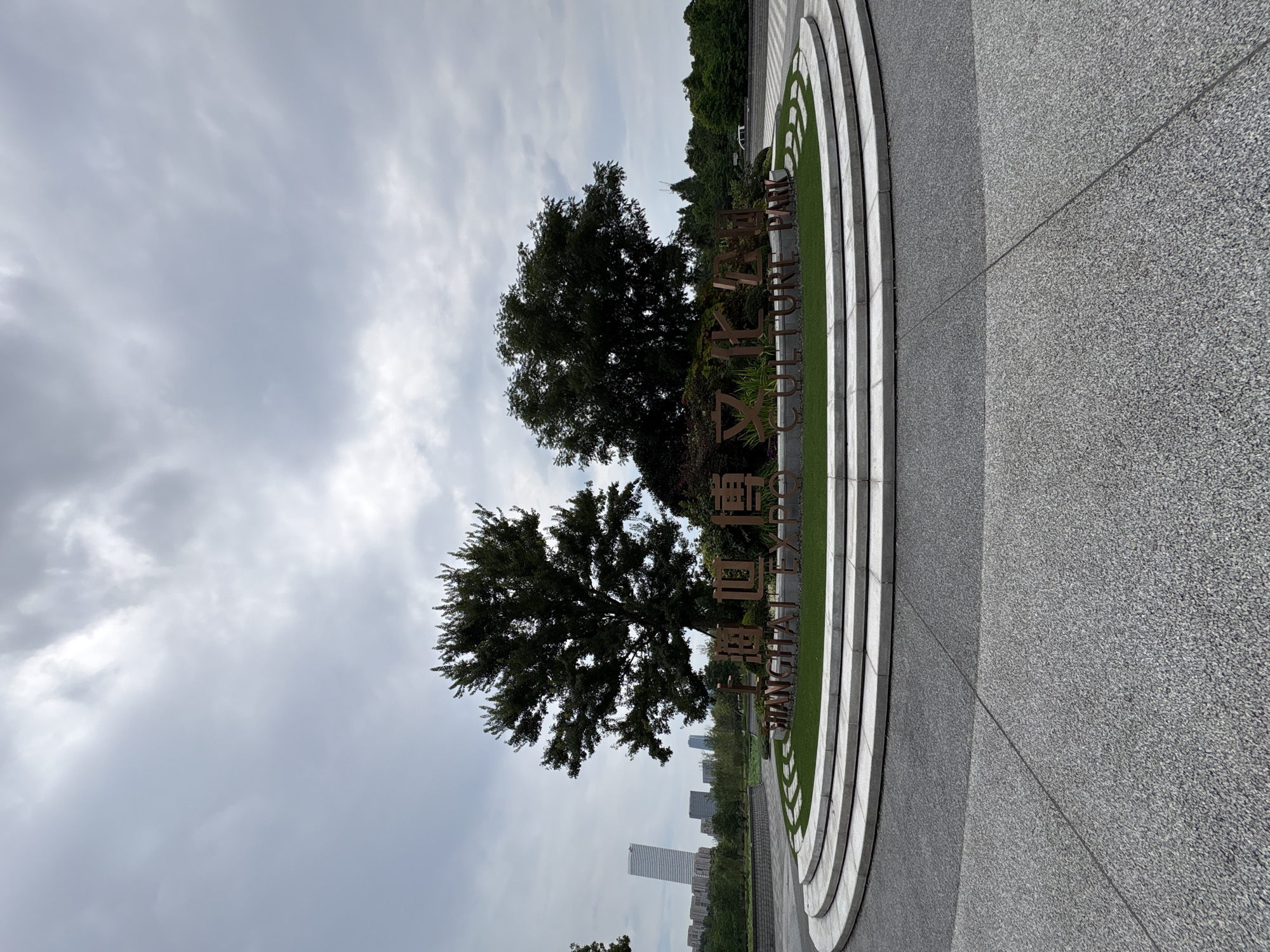 The image shows the "SHANGHAI EXPO CULTURE PARK" sign in Pudong, China, under a cloudy sky. The sign is made of bronze-colored metal letters and is set against a backdrop of lush green grass and trees. Two large trees are prominent in the foreground, with one on the left being larger and fuller than the one on the right. In the background, a modern skyscraper and other buildings are visible, indicating an urban setting. The sign is surrounded by tiered, circular concrete structures with grass inlays. The ground in front of the sign is paved with grey aggregate. The overall scene is one of a well-maintained park or public space.