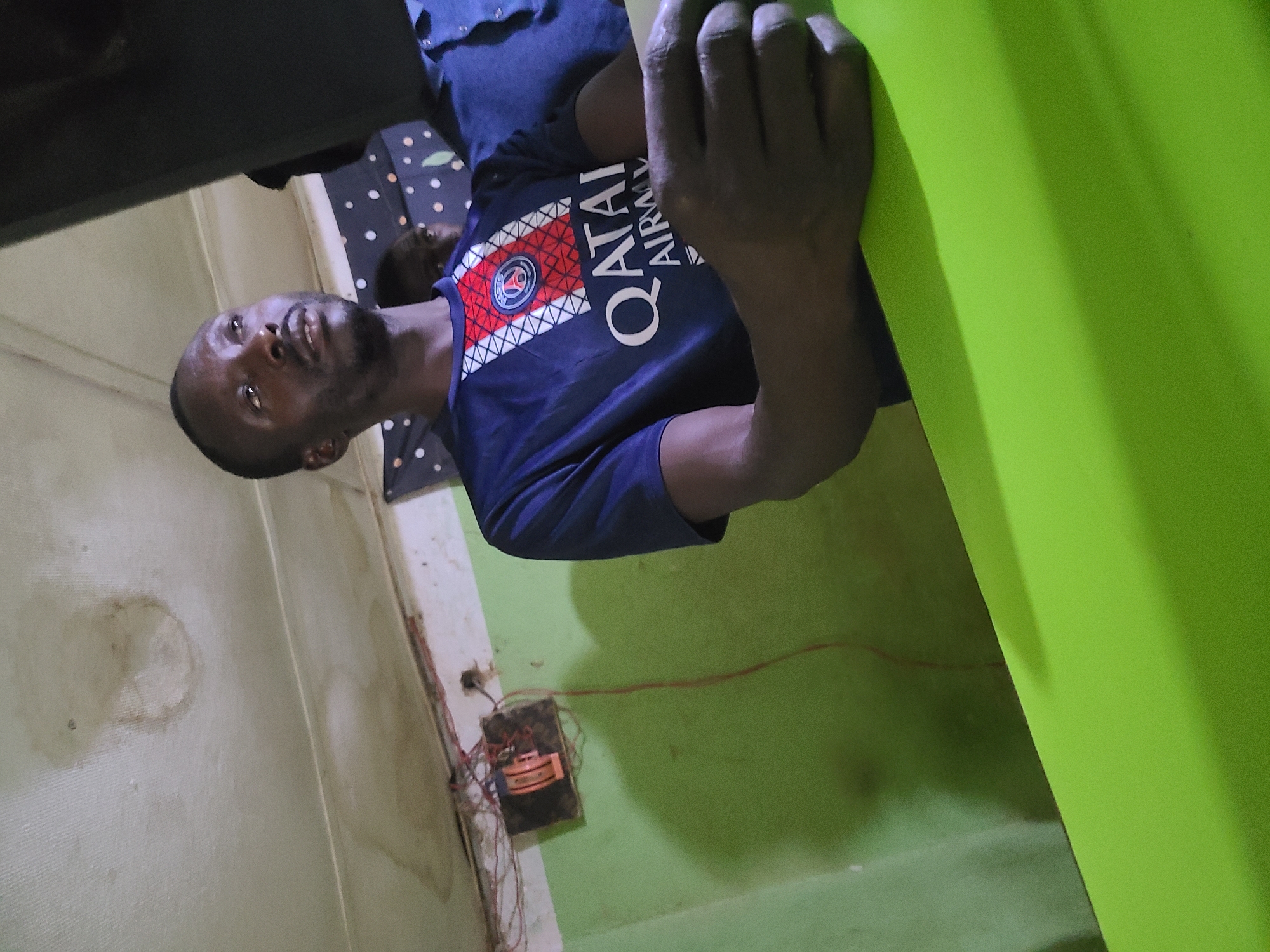 A man is lying on a bed in a room with lime green walls and a weathered white wall. The man has dark skin and a beard, and he is wearing a blue Paris Saint-Germain t-shirt. A dark, unidentifiable object is located in the top left corner. The room has an exposed electrical box on the white wall and an orange extension cord running along the green wall. The scene may be located in Jos, Nigeria, based on the user's request.