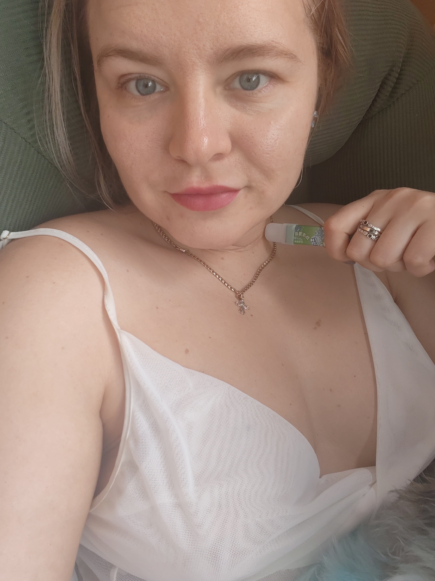 A close-up, high-angle selfie shows a fair-skinned woman with light blonde hair and blue eyes, wearing a white dress with thin straps. She has on a necklace with a small pendant and multiple rings on her right hand, which is holding a small, white tube with green writing. She is resting against a green, textured surface, possibly a chair or cushion. The overall lighting suggests an indoor setting. There is no indication of the location being Izhevsk, Russia.