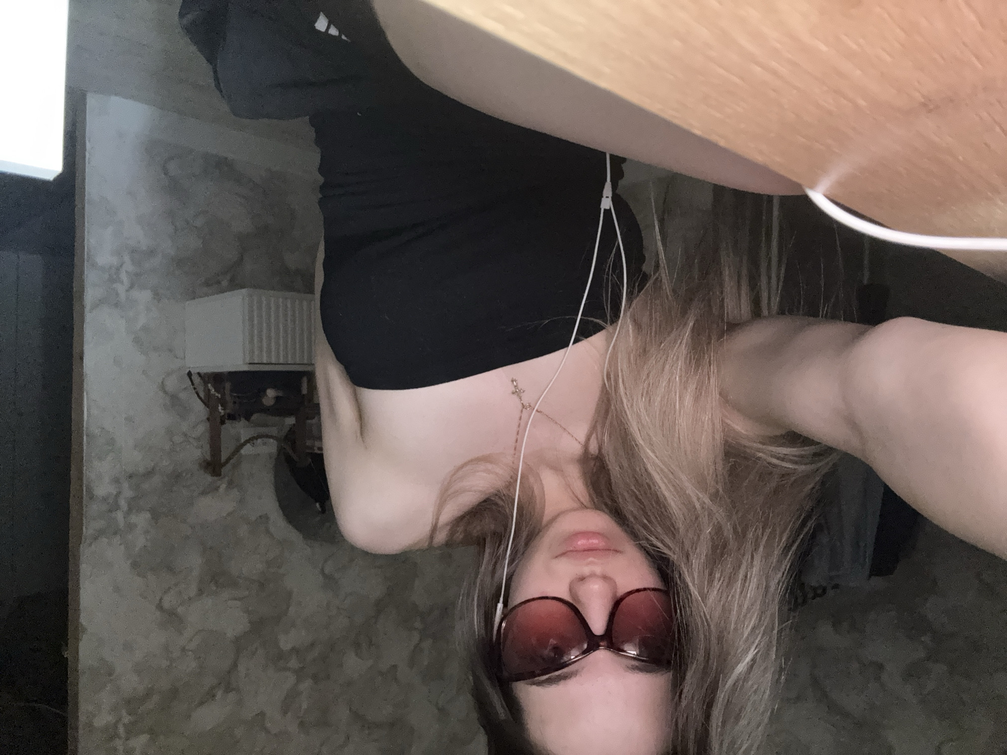 A woman is seen from a low angle, appearing to be taking a selfie. She is wearing sunglasses and a black top. She also has headphones in her ears and is wearing a necklace with a cross pendant. A wooden desk or table edge is visible above her. The background suggests an interior space with textured walls. There is a radiator and pipes visible on the wall in the lower left corner. The location of this image in Noviy, Russia cannot be verified.