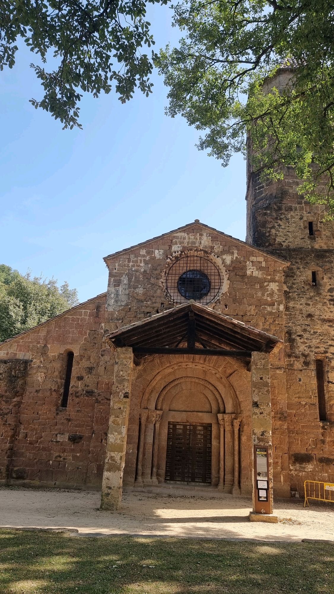 The image displays the stone facade of an ancient church in Sant Joan les Fonts, Spain. The building, constructed from rough-hewn, reddish-brown stone blocks, exhibits signs of age and weathering. A prominent feature is the arched doorway, topped with a semicircular tympanum and flanked by carved columns. Above the entrance, a wooden canopy juts out, supported by stone pillars. A large, circular window with a metal grate is visible high on the facade. To the right of the main building, a tall, cylindrical bell tower rises, also constructed of stone. The surrounding environment includes trees with green foliage and a clear blue sky. The ground in front of the church is a mixture of gravel and grass. A yellow barrier and an information sign are present near the entrance.