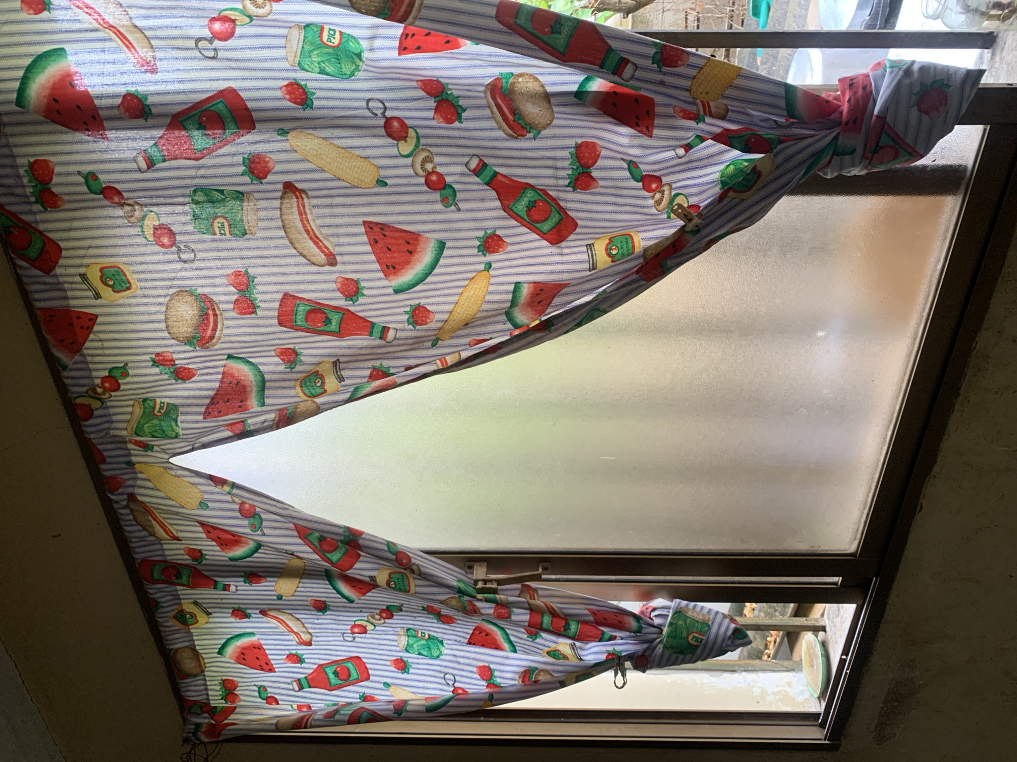 The image captures an interior view looking out through a window partially obscured by a patterned curtain. The curtain, featuring blue and white stripes, is adorned with various food illustrations commonly associated with picnics or barbecues. Notable items on the fabric include slices of watermelon, whole strawberries, corn on the cob, hot dogs in buns, hamburgers, bananas, and skewers loaded with vegetables like mushrooms and cherry tomatoes. Several condiment containers are also depicted, with "TOMATO" and "KETCHUP" visible on bottles of ketchup, and "PICK" clearly legible on jars or cans of pickles.
The curtain is tied back on both the upper and lower sides of the window frame, creating an opening in the center. The window itself has a metal frame; the upper pane is made of frosted glass, while the lower pane is clear. Through the clear lower section, outdoor elements such as green foliage and indistinct structures can be glimpsed, suggesting a view into a garden or courtyard. The lighting indicates it is daytime, with light filtering through the window, but the scene is not intensely bright. No human activity is taking place. The setting is an indoor space in Santa Ana, Venezuela.