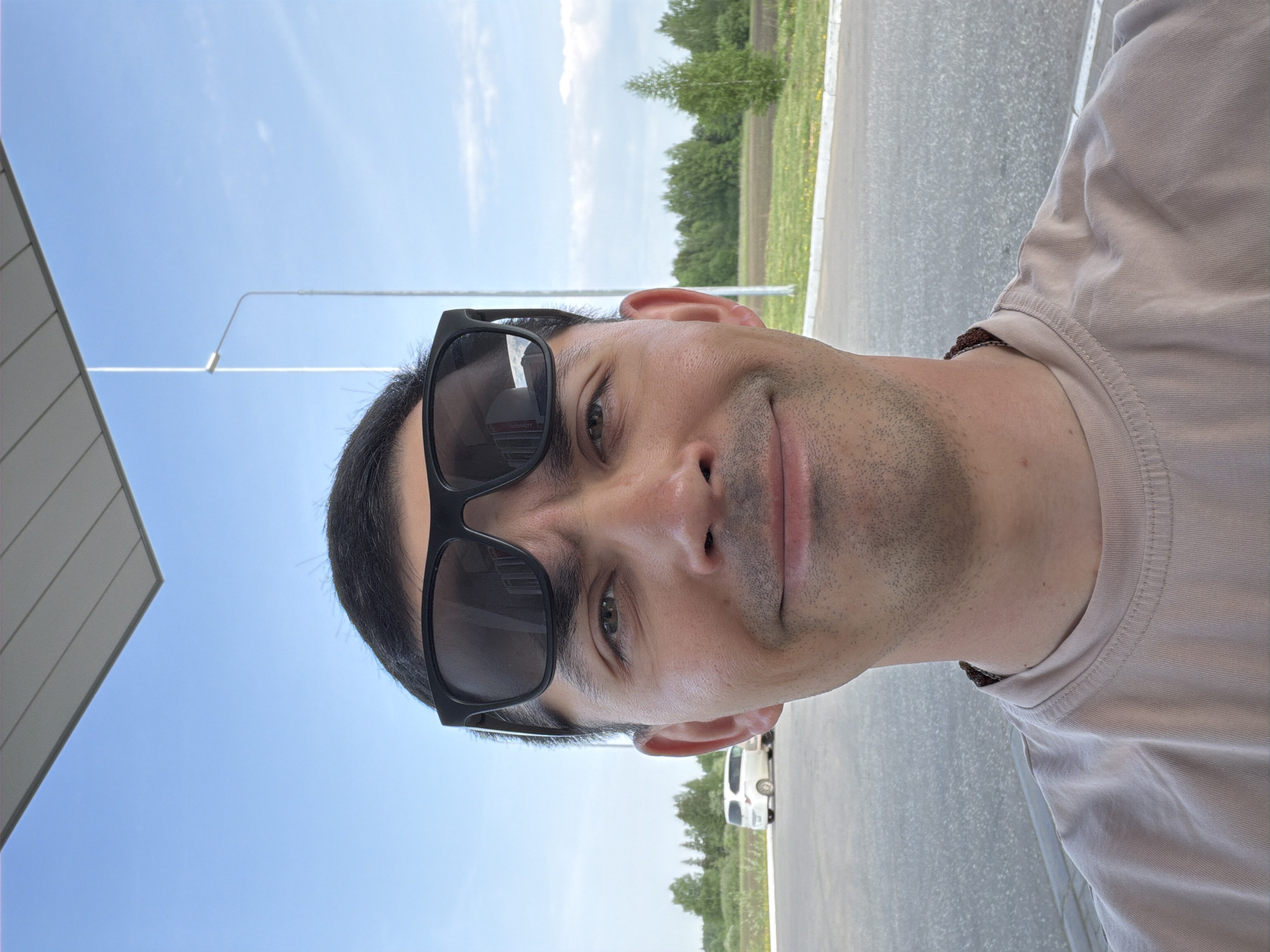 A man wearing sunglasses is shown in a close-up, tilted angle shot. The background includes a blue sky, a street light, a building, and a road with a car parked beside it. The surrounding area appears to be a green field with trees. The location is Unknown, Unknown.