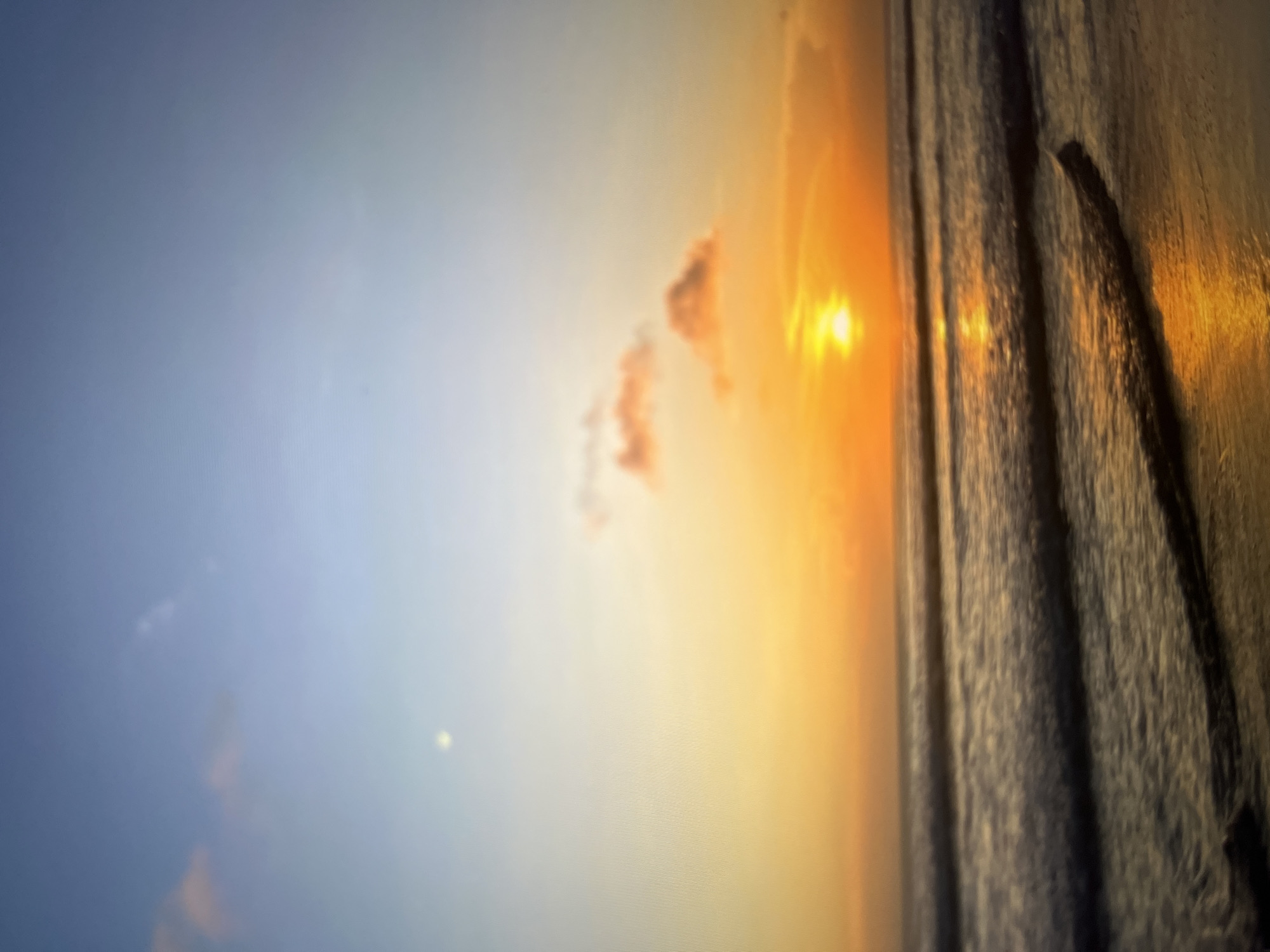 The image displays a tranquil beach scene at sunset. The sky transitions from a pale blue at the top to a warm orange and yellow gradient near the horizon, indicative of the setting sun. Several wispy clouds are scattered across the sky, tinged with pink and orange hues from the sunlight. The sun itself is a bright, round orb positioned low on the horizon, casting a golden reflection onto the water. Gentle waves roll towards the sandy shore, their surfaces shimmering with the light. The foreground consists of wet sand, with a dark, wet line marking the high tide line. The image is characterized by its soft focus and the warm, atmospheric lighting. The specific location in Cirahayu, Indonesia, is a coastal area. No people or specific objects are visible beyond the natural elements of the beach, ocean, and sky.