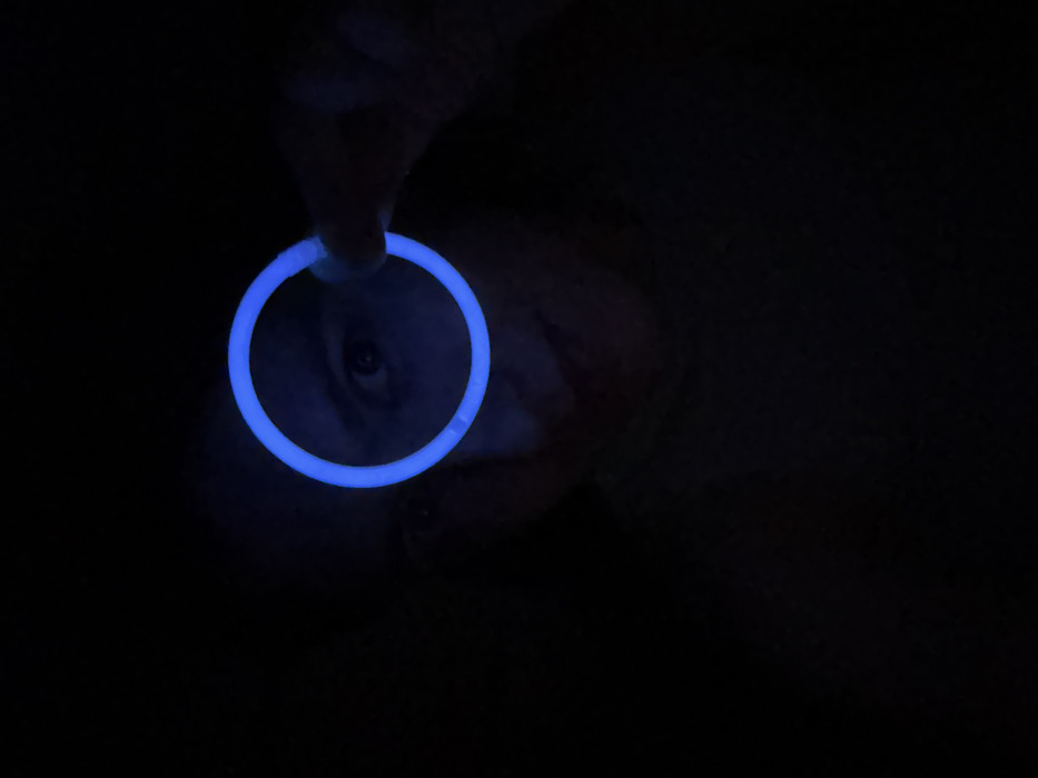 In near total darkness, a bright blue glowstick circle dominates the left side of the frame. A hand holds the glowstick, with fingers barely visible. An eye peers through the circle. Faint facial features are discernable behind the light, hinting at a person's face in the darkness. The rest of the image fades into deep shadow.