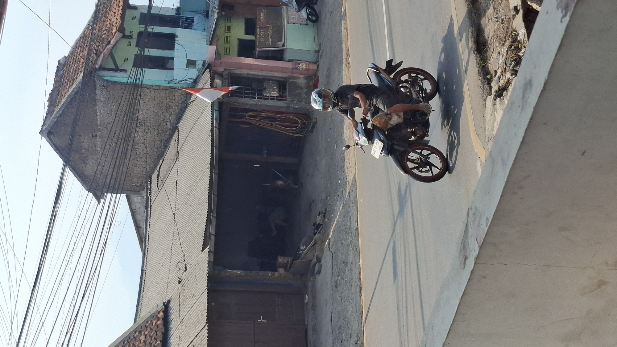 A man wearing a helmet rides a motorcycle on a paved road in Cilaku, Indonesia. The motorcycle is black with red accents on the wheels. The man is wearing a dark t-shirt and shorts. In the background, there are several buildings with corrugated metal roofs, and a flag of Indonesia is visible. Power lines are strung across the sky. The scene appears to be a street in a residential or commercial area.