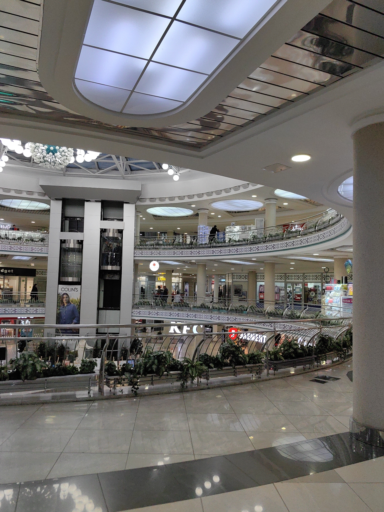The image shows an interior view of the Galleria Minsk shopping mall, located on Prospekt Pobediteley in Minsk, Belarus. The scene captures multiple levels of the mall's interior, characterized by a modern, multi-story design with escalators, elevators, shops, and open walkways.
In the foreground, a polished tiled floor reflects the ambient light and the mall's architecture. A dark gray band runs across the floor, dividing it into sections. A metallic railing with a decorative pattern separates this area from a lower level.
The midground reveals a central elevator tower with glass-paneled elevators. On the side of the tower, a large advertisement for "COLIN'S" clothing is displayed, featuring a model. Several levels of walkways and shopfronts are visible on either side of the elevator shaft, adorned with railings. Plant displays are integrated along the edges of the walkways. Some shop names are partially visible, such as "BUDGET" and "KFC".
The upper levels showcase rounded balconies and more shopfronts, with people browsing and walking. The ceiling features a large, modern lighting system with translucent panels and decorative suspended lights, creating a bright and airy atmosphere. The overall impression is one of a bustling commercial space with an emphasis on contemporary design and an abundance of natural and artificial light.