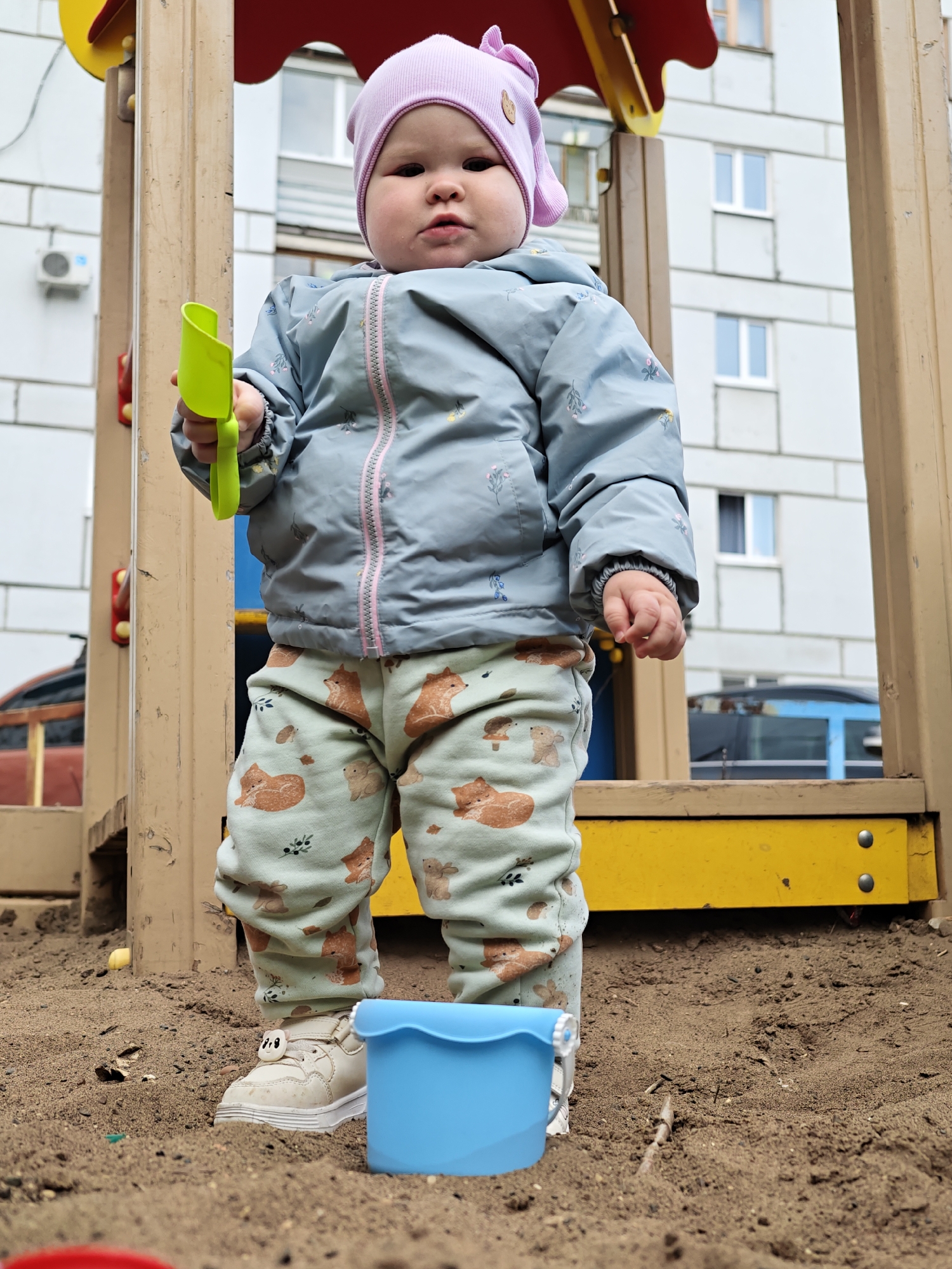A toddler stands in a sandbox within a playground structure in what appears to be an urban setting. The child wears a light blue patterned jacket, animal-print pants, a pink hat, and dirty off-white shoes. The toddler holds a green plastic shovel in their right hand and a blue plastic bucket is near their left foot. The sandbox is surrounded by a wooden play structure. In the background, an apartment building with white walls and gray-framed windows is visible. The scene appears to be located in Chaikovsky, Russia, based on the metadata associated with the image file.