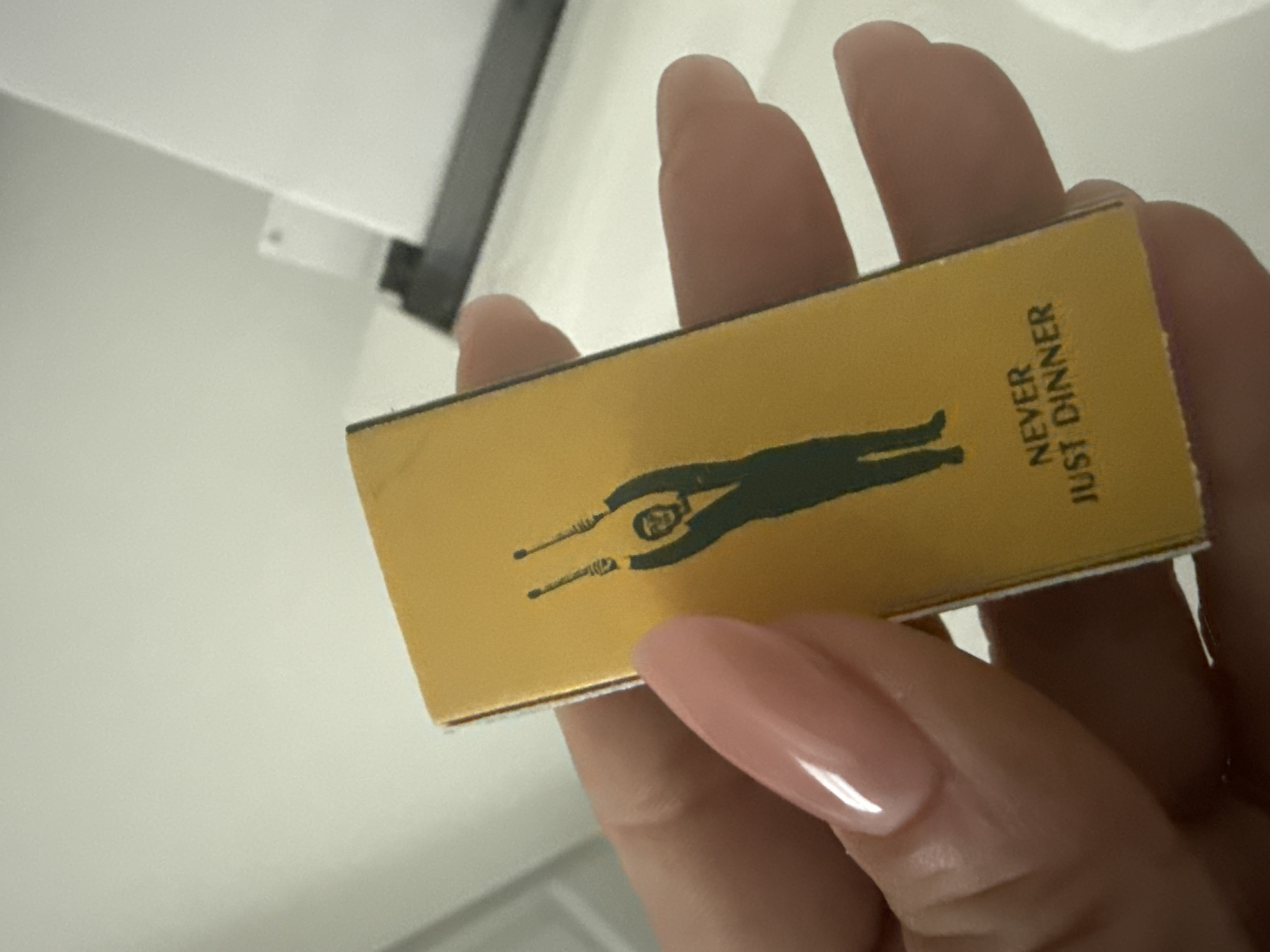 A close-up, slightly angled photograph captures a human hand holding a small, golden-yellow rectangular object, likely a matchbox or small container. The hand features light pink, almond-shaped, polished fingernails.
The held object is predominantly golden yellow with a distinct black graphic and text. The graphic is a silhouette of a person, possibly a man, standing with arms stretched upwards, holding two thin, stick-like objects. The figure's head displays some internal detailing. Vertically oriented on the right side of the object, the black text "NEVER JUST DINNER" is clearly visible.
The background is a bright, plain white or off-white surface, suggesting an indoor setting. A dark, linear element, possibly a shelf edge or a fixture, is partially visible in the upper left portion of the image. The lighting is bright and even, characteristic of an interior space. No specific activity beyond holding the object is depicted, and no emotions are discernible. The image provides no specific location cues for Davie, United States of America.
