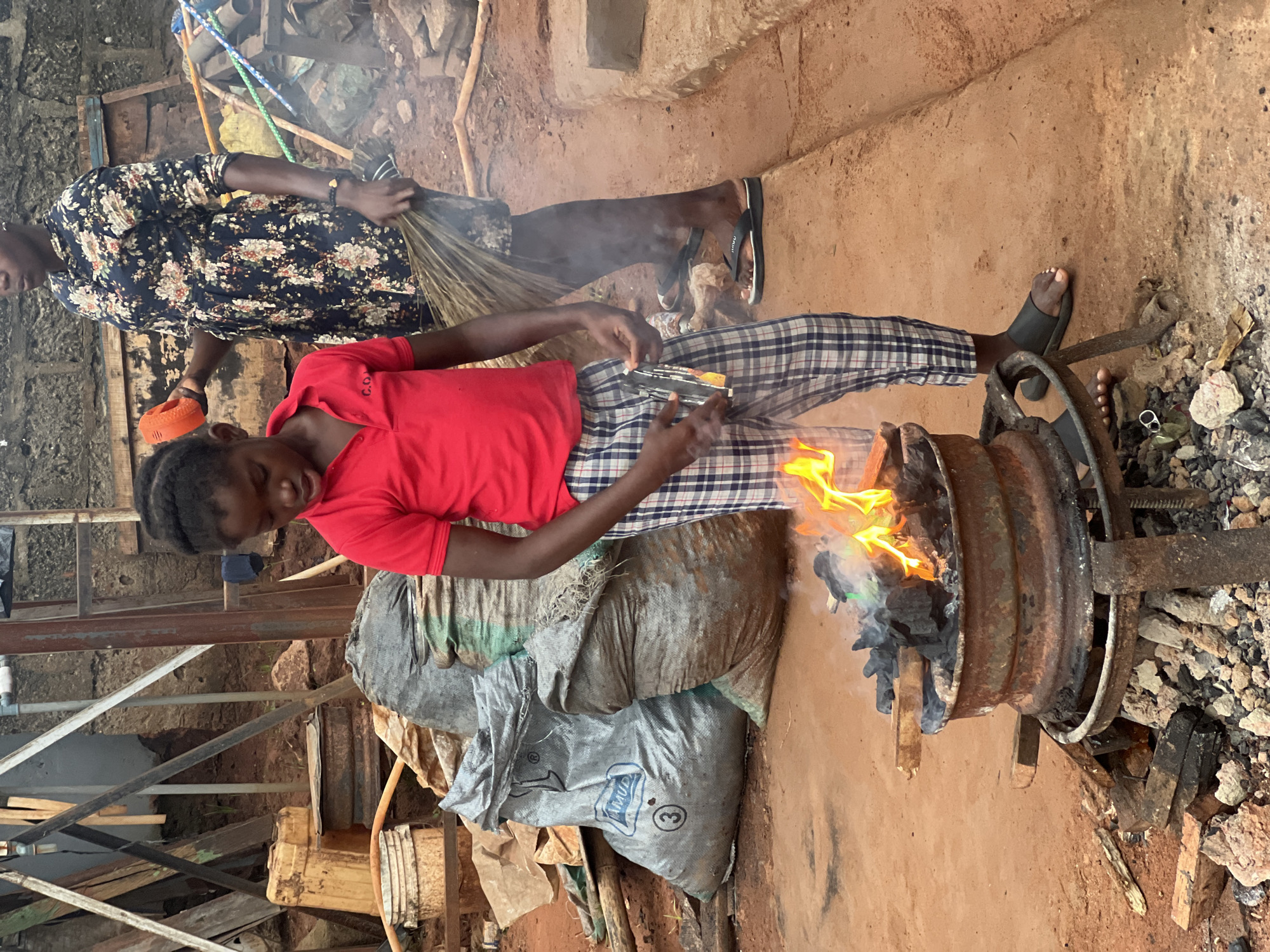 The image depicts two individuals in an outdoor setting, likely a workshop or yard, in Nkpor, Nigeria. The scene is characterized by a dirt ground and what appears to be a traditional furnace or forge. In the foreground, a young person wearing a red t-shirt and plaid pants is focused on a metal drum filled with burning charcoal. Flames erupt from the drum, indicating an active process of heating or smelting. The individual is holding what looks like metal rods or tools, suggesting they are actively engaged in metalwork.
Behind this individual, another person, dressed in a dark, floral-patterned shirt, stands and holds a broom, appearing to be sweeping or preparing to sweep. Their posture suggests they might be involved in maintaining the workspace.
The environment is rustic, with visible piles of sacks, possibly containing materials for the work, and scattered debris. The lighting suggests it is daytime, likely mid-morning or afternoon, with no distinct weather conditions apparent. The overall atmosphere is one of active labor and traditional craftsmanship.
There is no visible text in the image that is legible. The focus is on the manual labor and the primitive yet functional tools and setting.