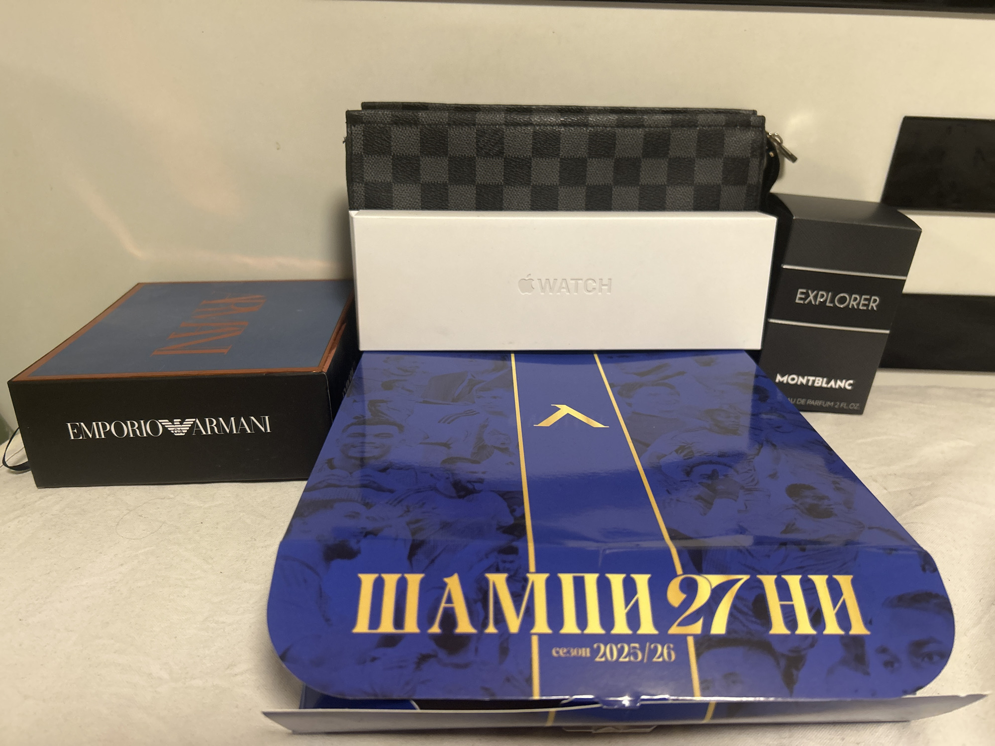 This media file presents a collection of luxury and branded items arranged on a light-colored surface, likely indoors in Dunavtsi, Bulgaria. The background is a plain light wall.
The objects visible include:
* A black and grey checkered patterned wallet or small pouch, resembling the Louis Vuitton Damier Graphite pattern, positioned atop a white box.
* A white Apple Watch box with the "WATCH" logo prominently displayed.
* A black box for "MONTBLANC EXPLORER" fragrance, visible with "EAU DE PARFUM 2 FL. OZ.".
* A dark blue box with "EMPORIO ARMANI" logo in white, positioned on the left.
* A large blue box or flat item, possibly a book or game board, with gold Cyrillic text and faded images of people (likely athletes or celebrities) in the background. The text reads "ШАМПИОН 27 НИ" and "сезон 2025/26". The word "ШАМПИОН" translates to "CHAMPION".
The items appear to be new or recently acquired, given their packaging. The arrangement suggests they might be gifts, new purchases, or items being displayed. No human activity is depicted; the image is a still life of consumer goods. The lighting is even, suggesting an indoor setting without direct sunlight.