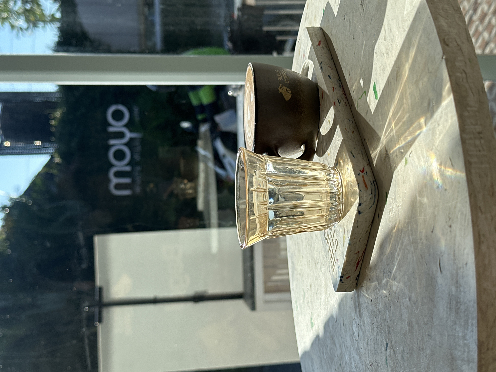 The photo depicts a partially visible coffee cup and an empty, ribbed glass resting on a weathered, possibly recycled material surface. Reflected in the window behind is the logo "moyo," suggesting the name of a cafe or establishment. The reflection suggests a location with foliage and possible activity, hinting at an urban setting. Considering the prompt's reference to North Kuta, Indonesia, it's likely this scene is within that vicinity, possibly in a cafe with outdoor seating reflecting the local environment. No people are directly visible, but the presence of the drinkware implies recent or intended use.