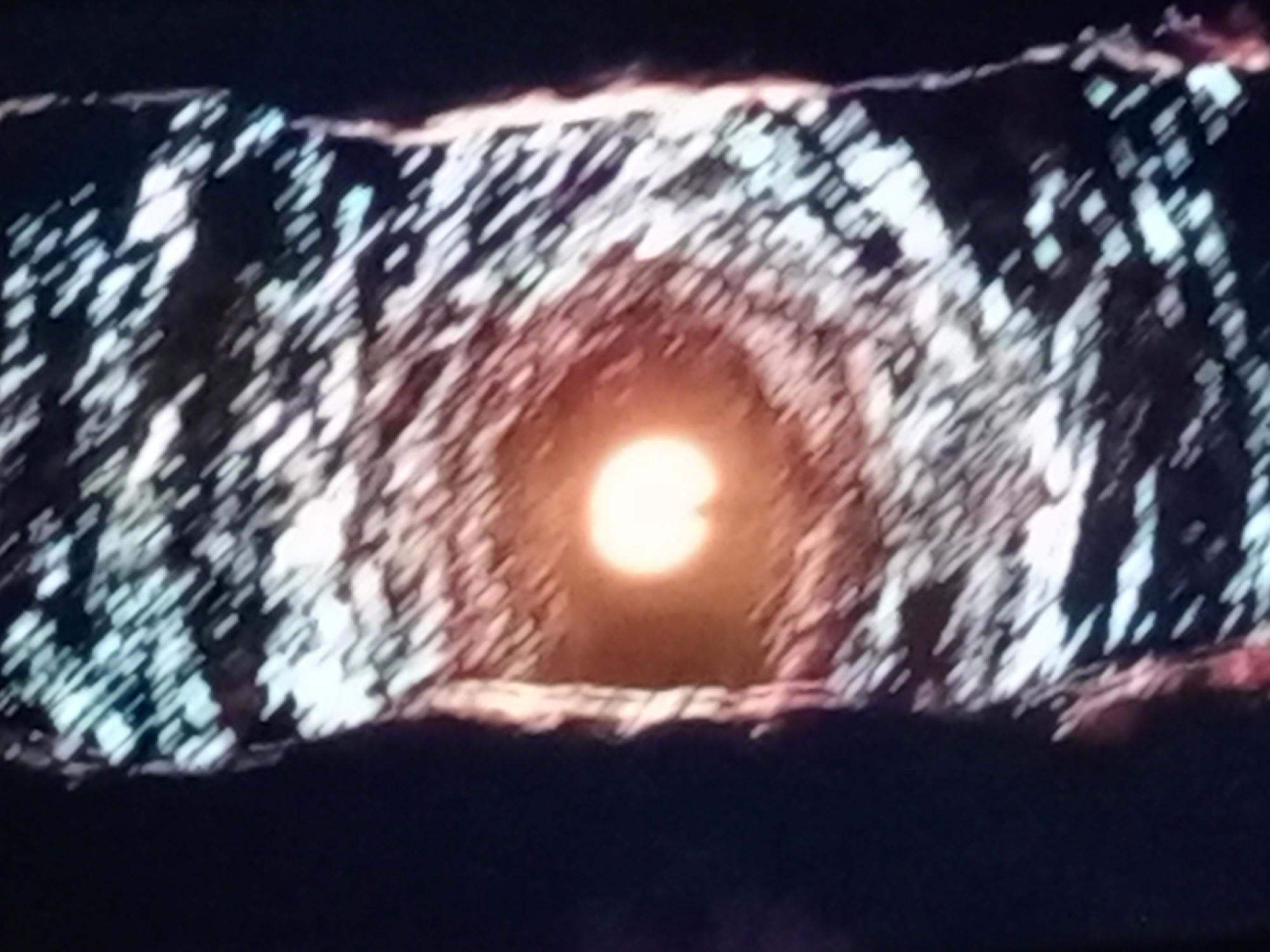 The image displays a tunnel-like structure with rough, textured walls that are illuminated by a bright, circular light source at its center. The walls appear to be a mix of light and dark shades, with diagonal lines of light suggesting a rough, possibly rocky or crystalline surface. The overall impression is of a natural cavern or an artificially created passage leading towards an intensely lit opening. The foreground and edges of the image are dark, creating a stark contrast with the illuminated interior. There are no discernible people or specific objects within the scene, and the environment is abstract and appears to be an unusual or otherworldly location.
