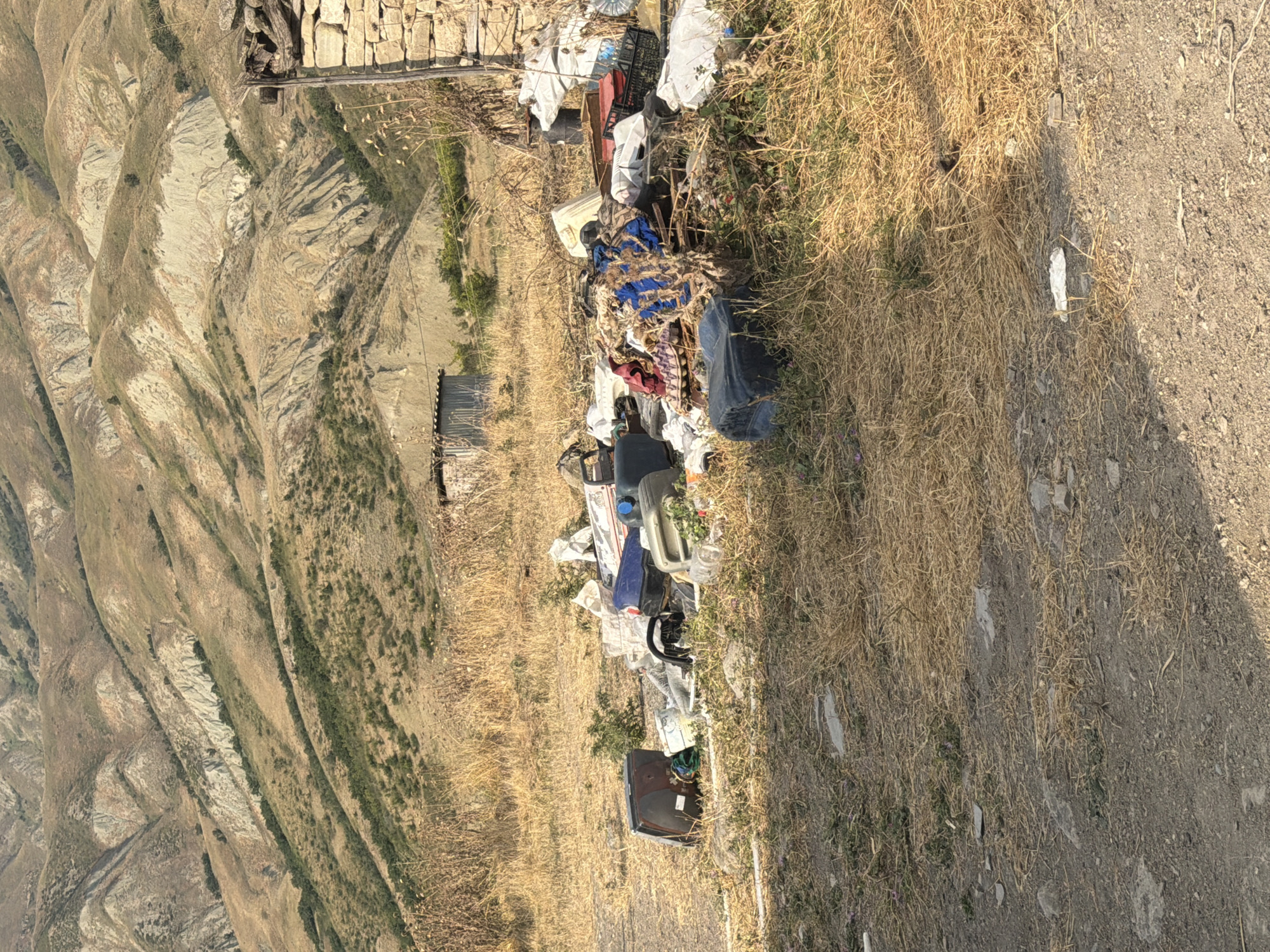The image depicts a pile of refuse, including plastic containers, bags, and assorted debris, situated in a dry, grassy area. In the background, a hillside with sparse vegetation and a dilapidated structure made of stone and corrugated metal is visible. The scene appears to be an outdoor setting in a rural or undeveloped area, likely in the vicinity of Köhnə Quşçu, Azerbaijan, given the arid landscape and rustic structures. There are no people or animals present in the image.